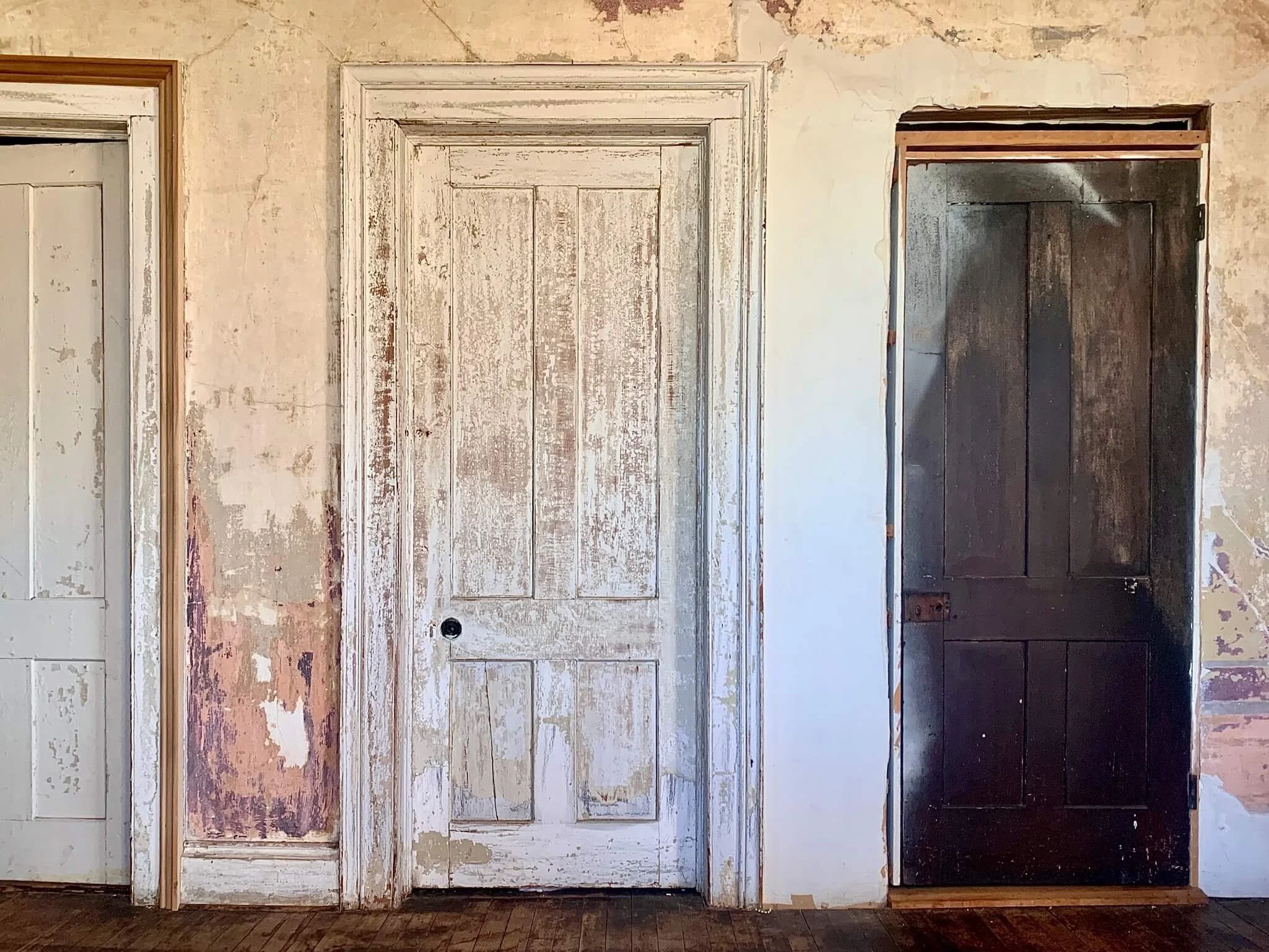 Three old wooden doors, with peeling paint, set in an aged wall with visible cracks and chips. The left door is painted white, the middle door is mostly white, and the right door is dark brown.