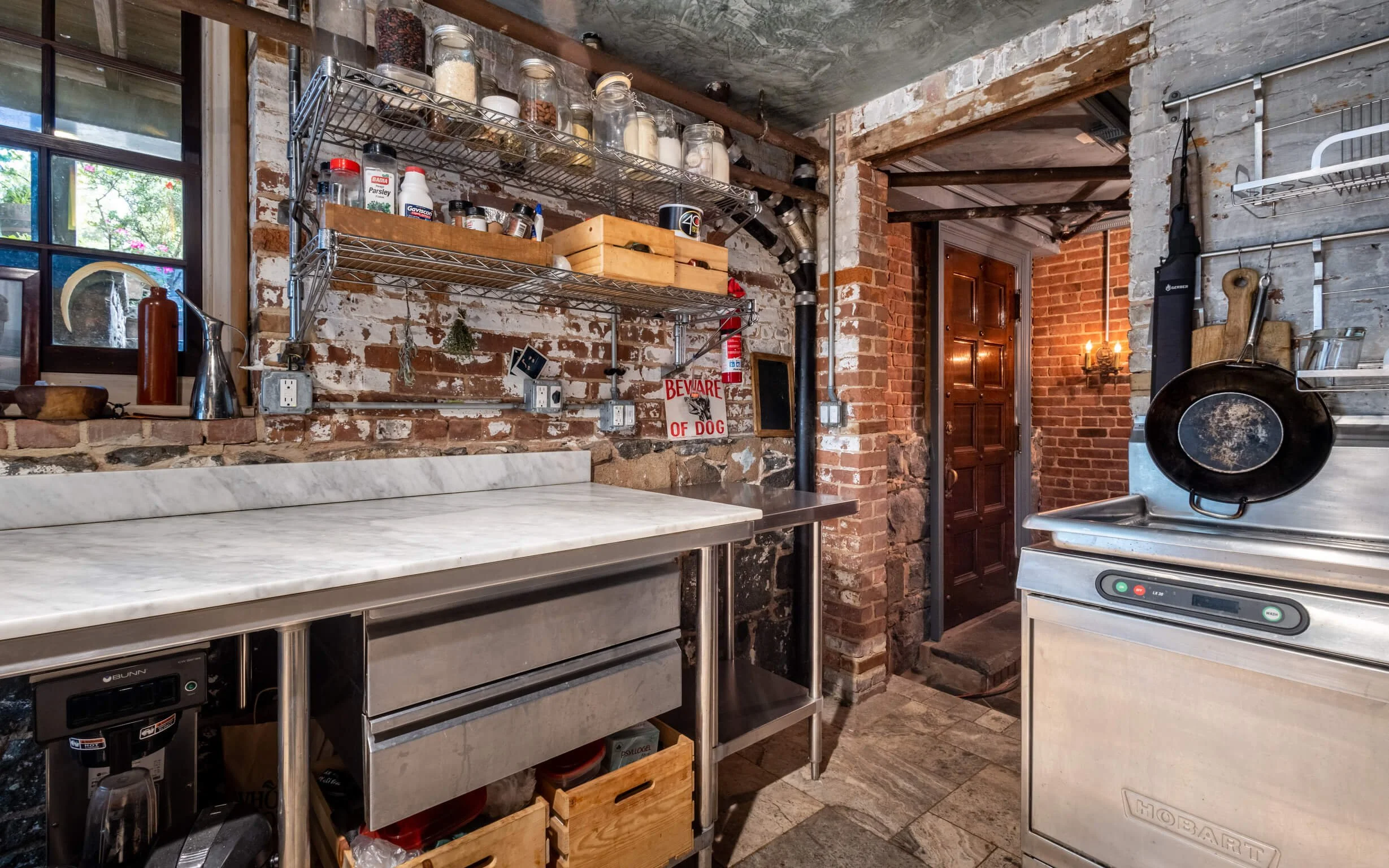 A rustic kitchen with exposed brick walls, a large marble countertop, open shelving with jars and containers, a window with a crescent moon decoration, and a metal stove with a cast iron skillet hanging on the side.