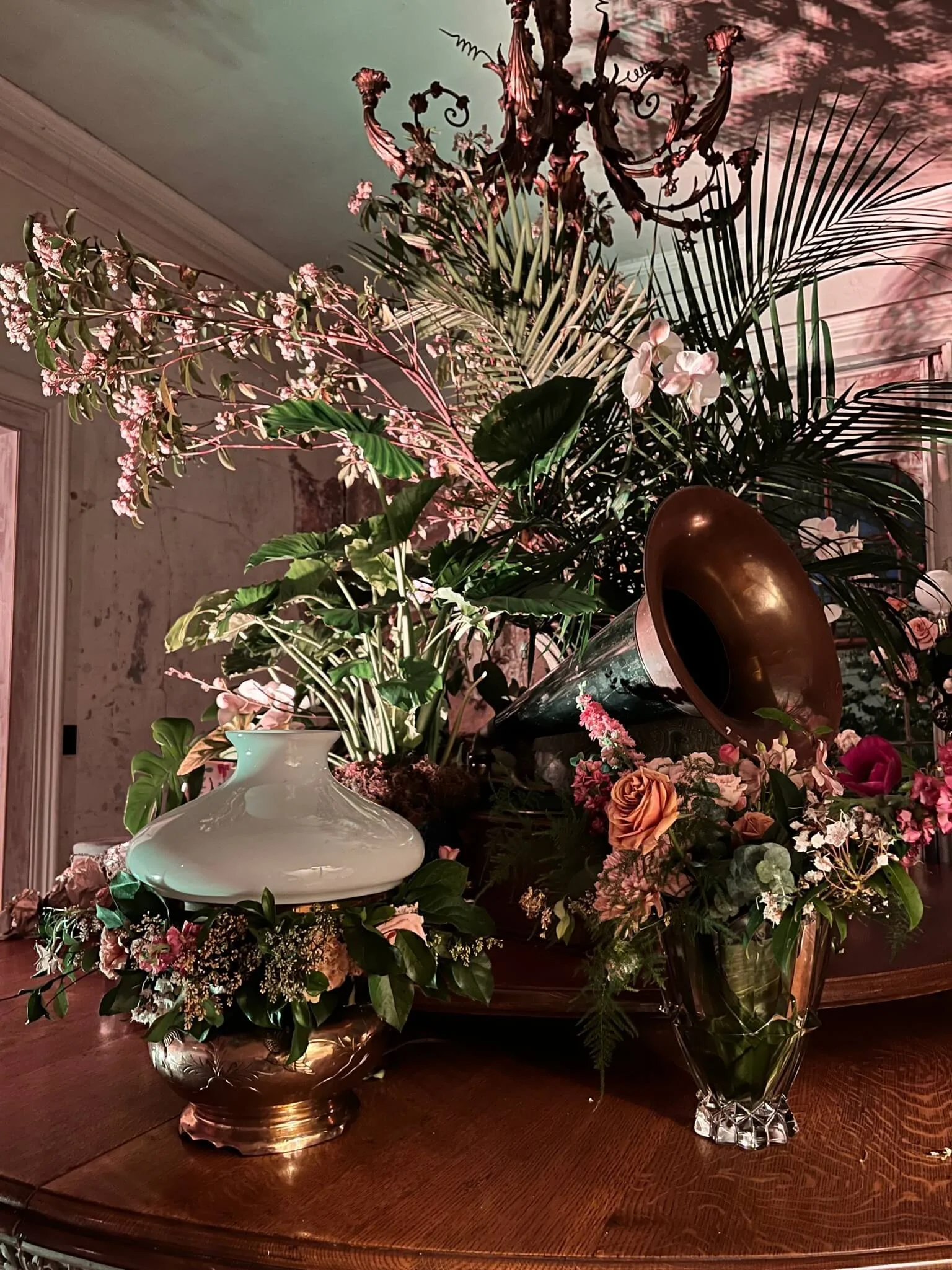 A floral arrangement on a wooden table with vases and a vintage gramophone, featuring pink and white flowers, large green leaves, and a copper-colored gramophone horn.