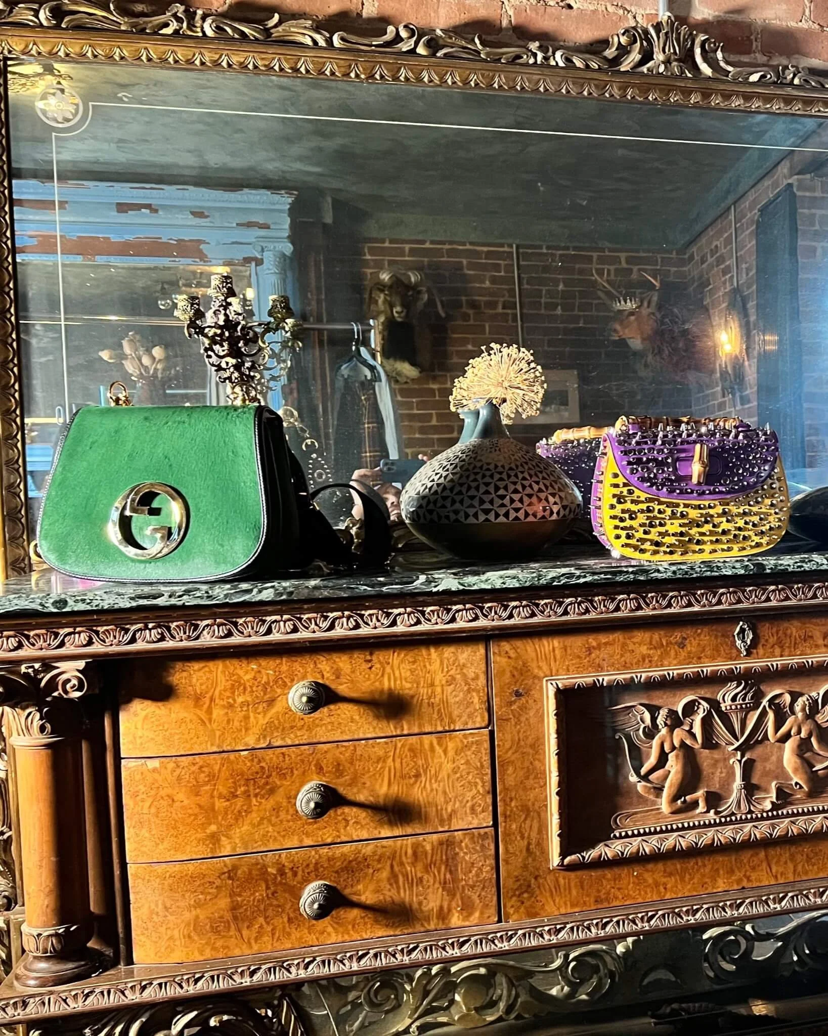 Decorative vintage wooden dresser with ornate carvings, topped with a large mirror and various fashion accessories like handbags and a vase with a dried flower, in a room with brick walls decorated with a mounted animal head and deer antlers.
