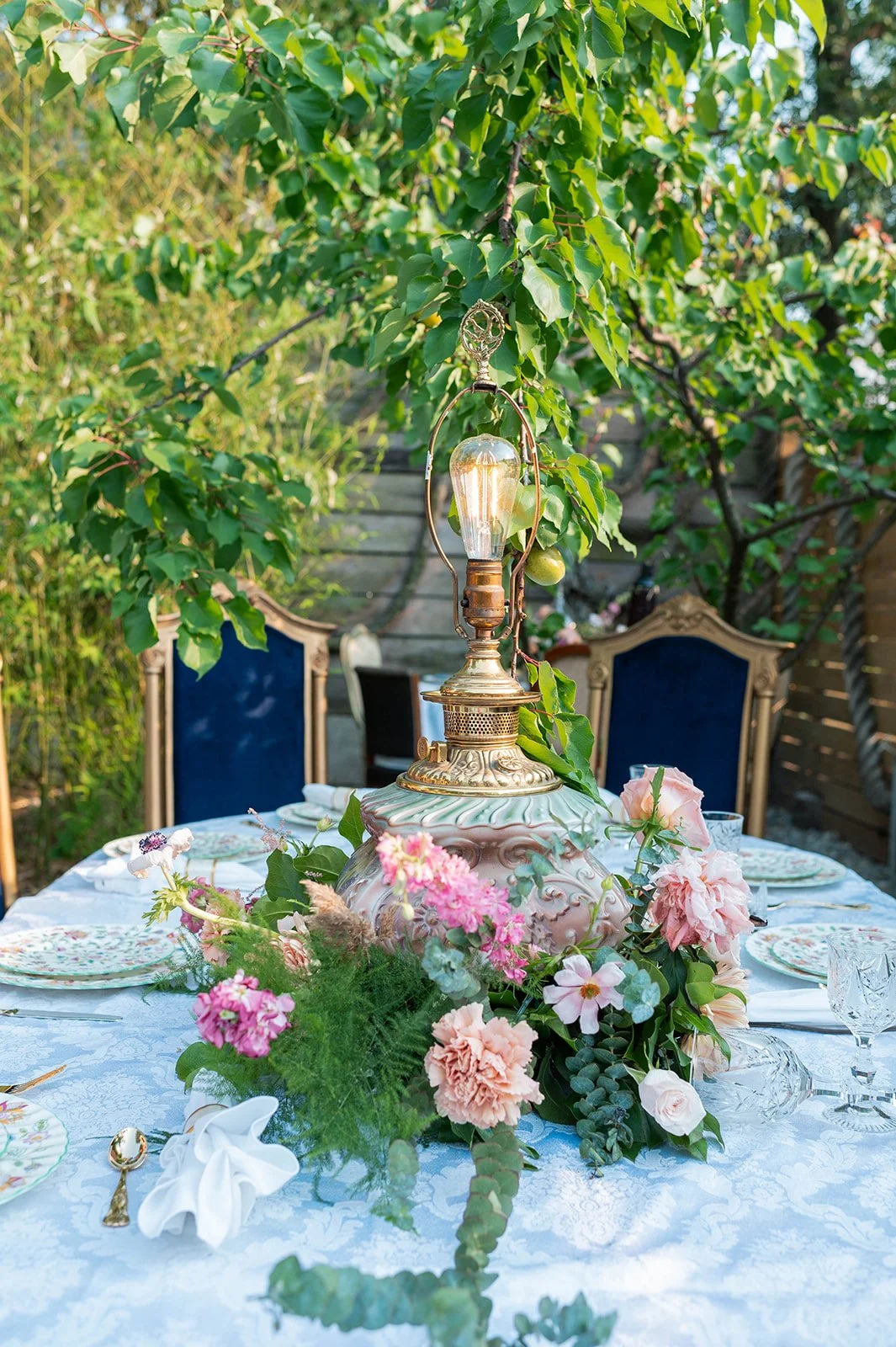 An outdoor dining table decorated with a floral centerpiece, vintage tableware, crystal glasses, and a decorative lamp with greenery in the background.