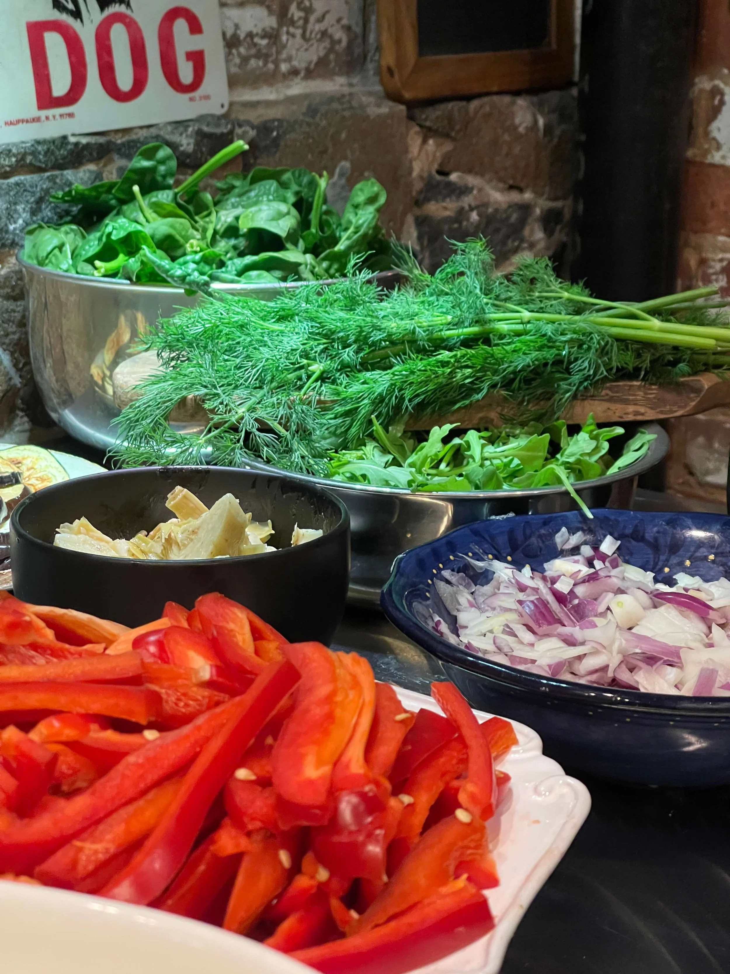 Assorted fresh vegetables including chopped red bell peppers, sliced onions, chopped artichokes, fresh green herbs, and leafy greens in bowls on a kitchen counter, with a brick wall background.
