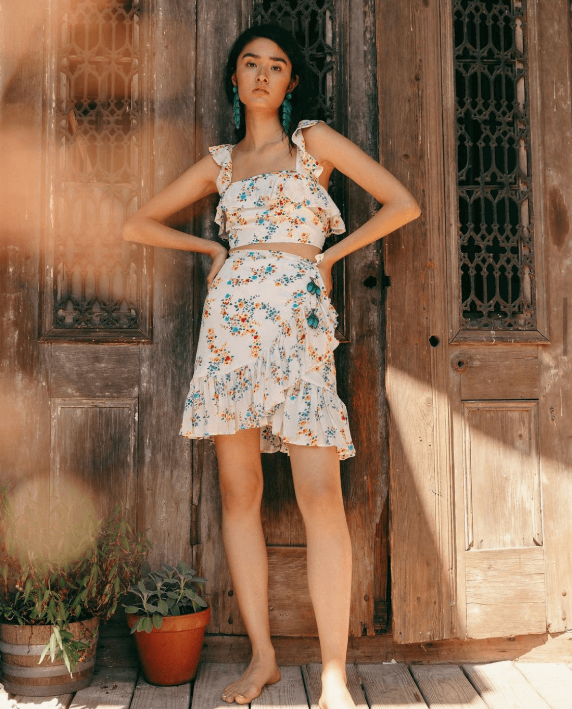 Young woman in floral summer outfit standing barefoot against a rustic wooden door, with potted plants nearby, on a sunlit outdoor porch.