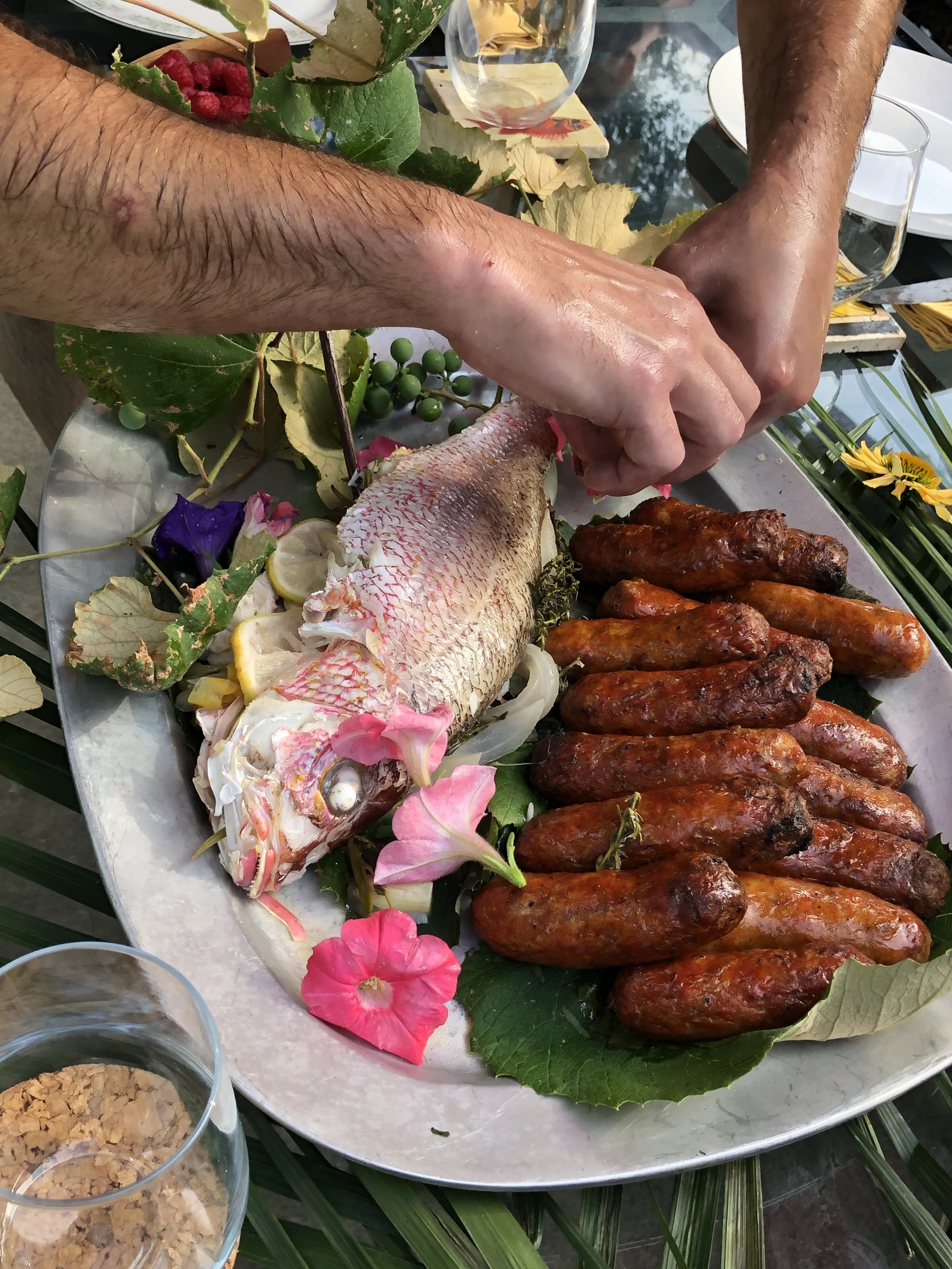 Person preparing a seafood dish with a whole fish and grilled sausages on a silver platter decorated with flowers and leaves.