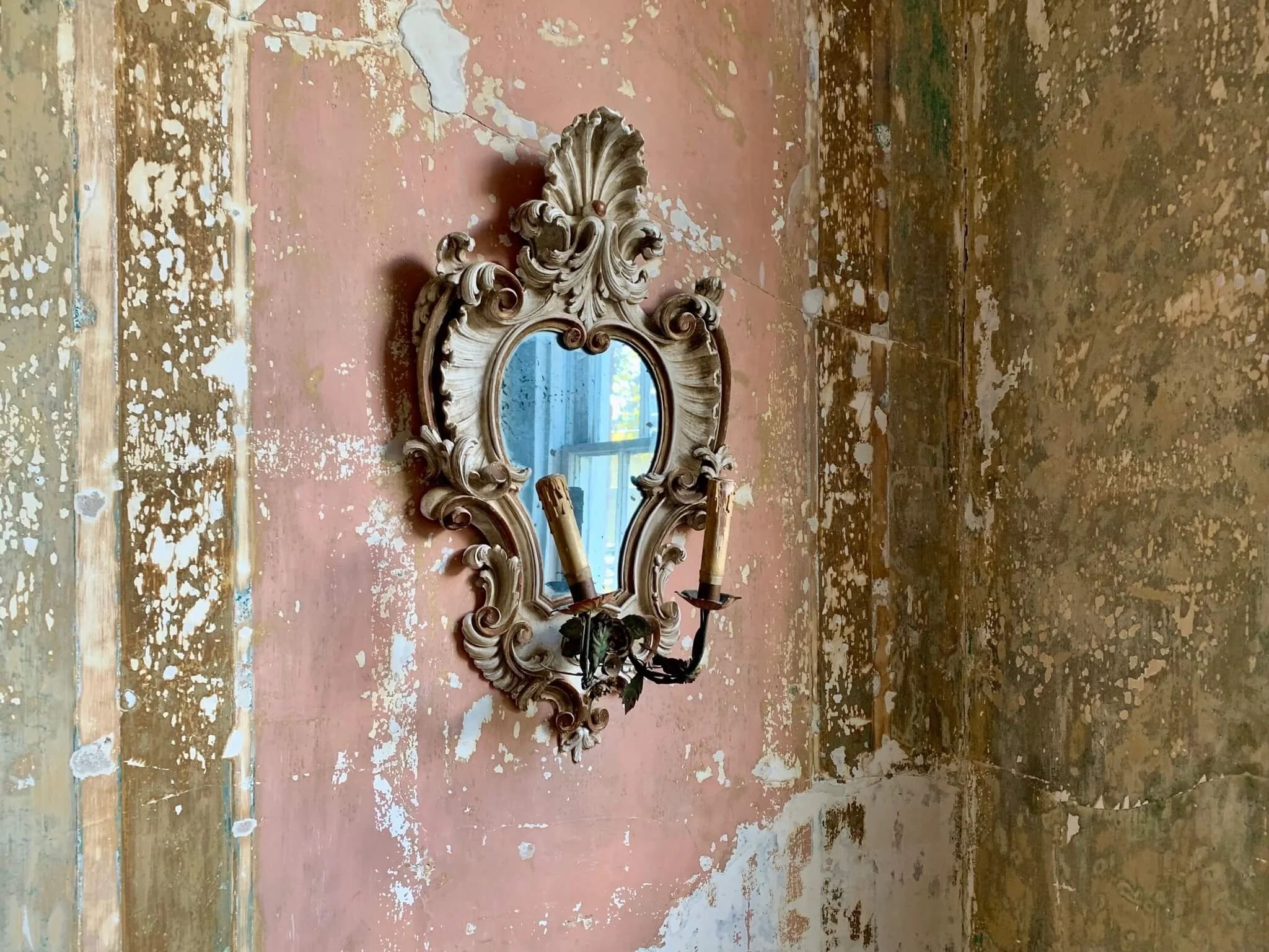 An ornate vintage wall mirror with a carved cream-colored frame, mounted on a worn, pink and beige wall in a rustic room. A black wrought iron candle holder with two candles is attached to the bottom of the mirror frame, with the candles missing thei