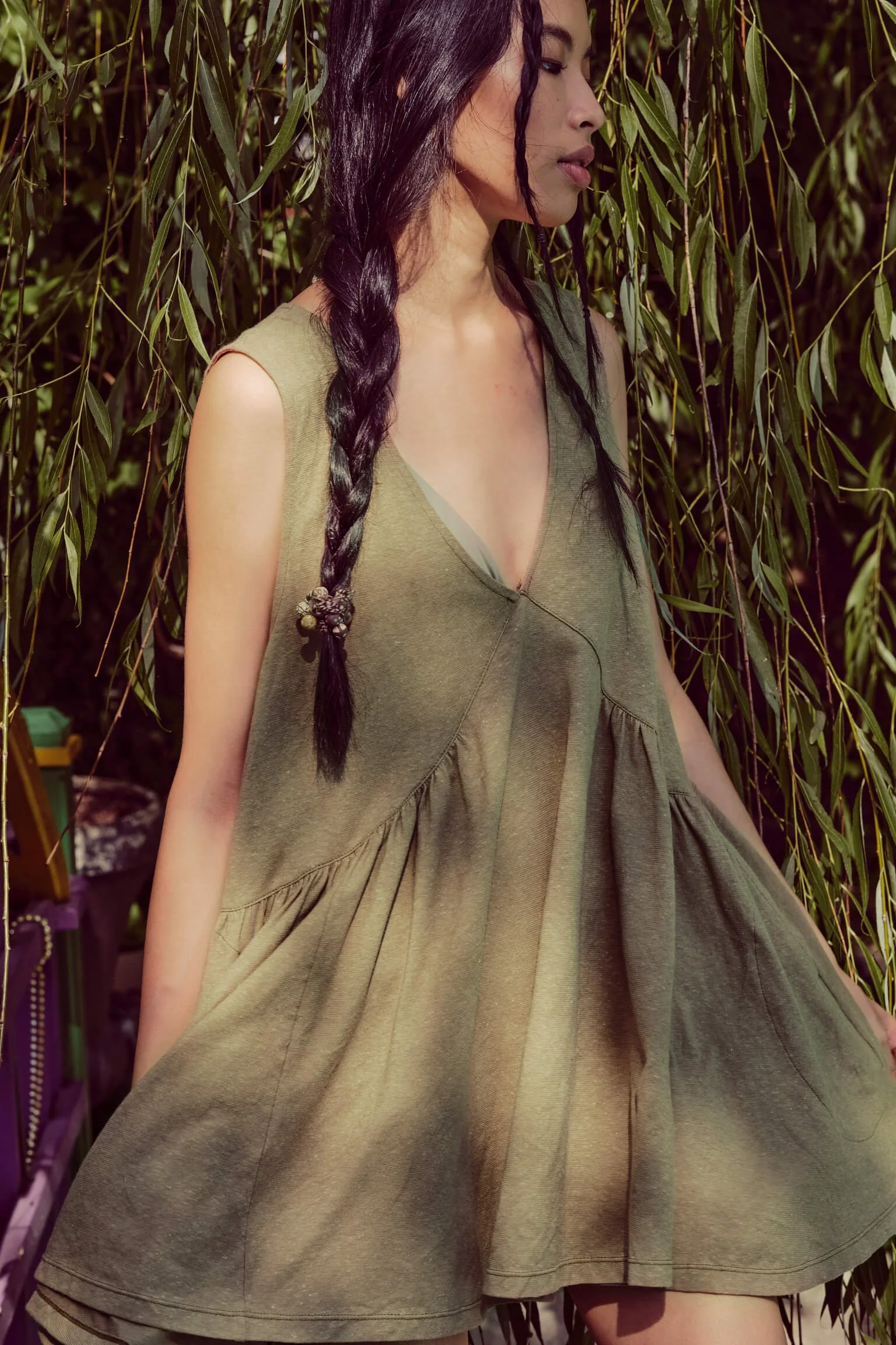 A woman with long dark braided hair standing outdoors near a tree, wearing a sleeveless, loose, olive-green dress with a deep V-neck.