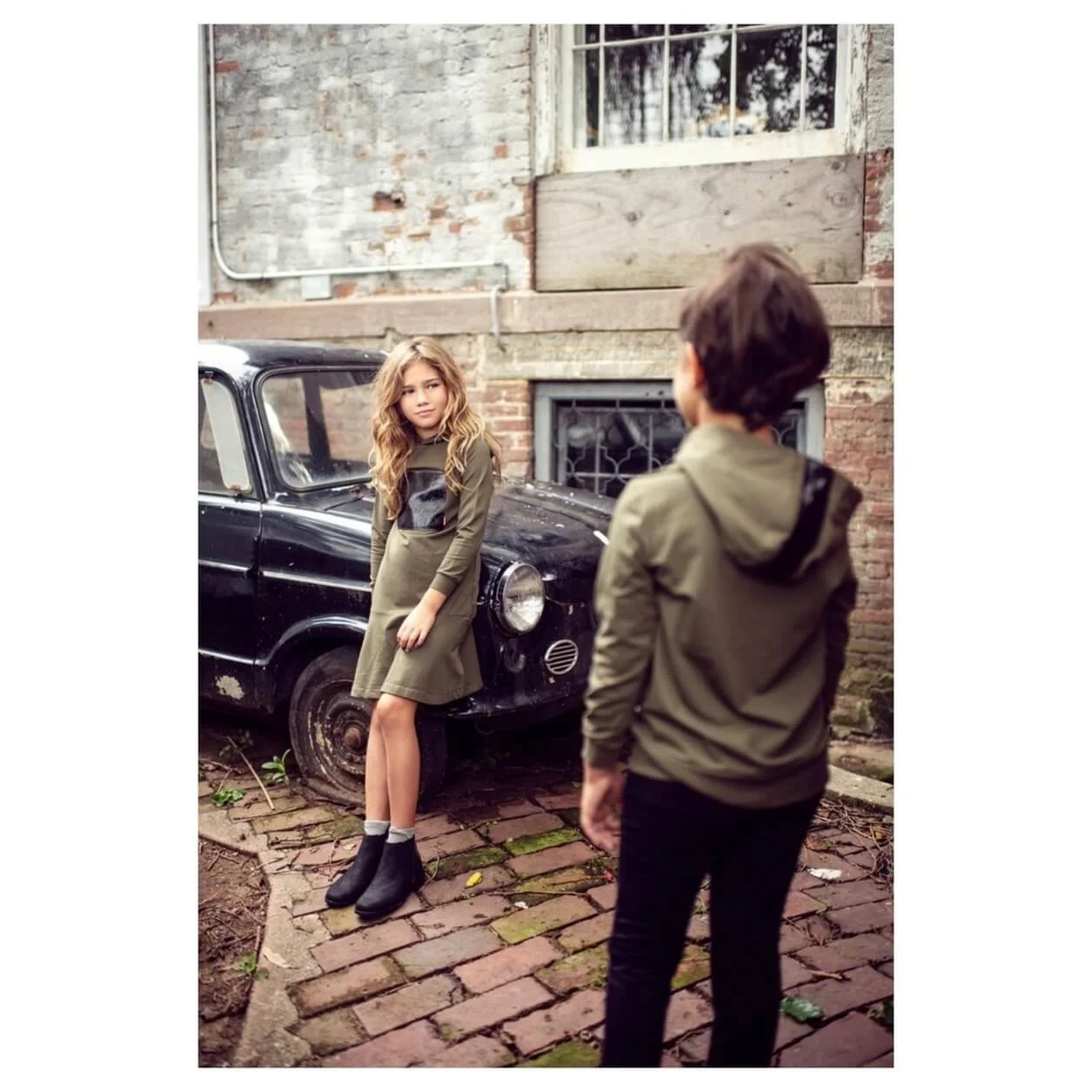 A girl with wavy red hair wearing a green dress and black boots leaning against an old black car, looking at a boy with brown hair wearing a green jacket, standing with his back to the camera in a brick yard.