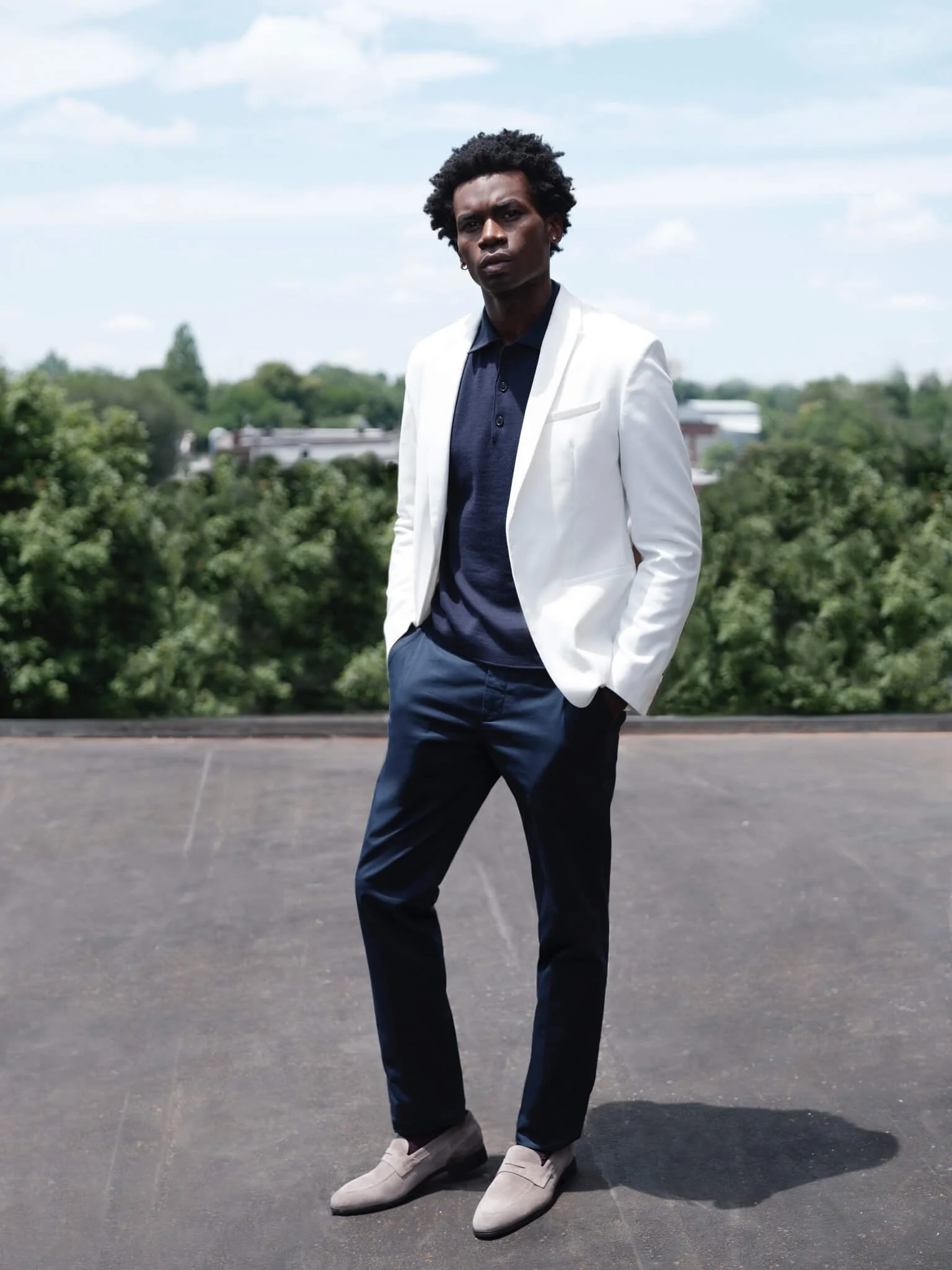 A young man standing outdoors on a paved surface with trees and a blue sky in the background, wearing a white blazer, navy shirt, navy pants, and light-colored shoes.