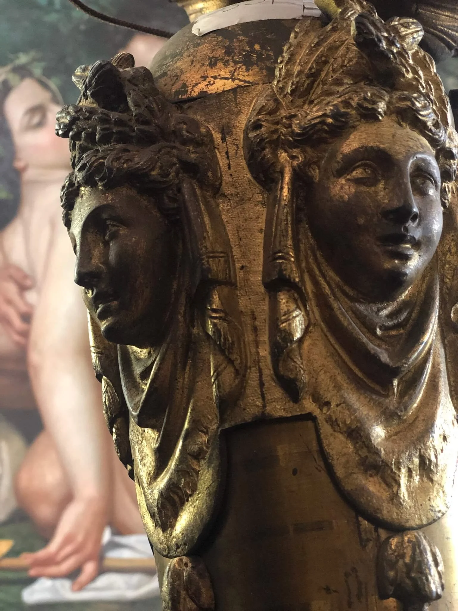Close-up of a wooden sculpture featuring two carved female faces with detailed hair and expressions, with a blurred person in the background.