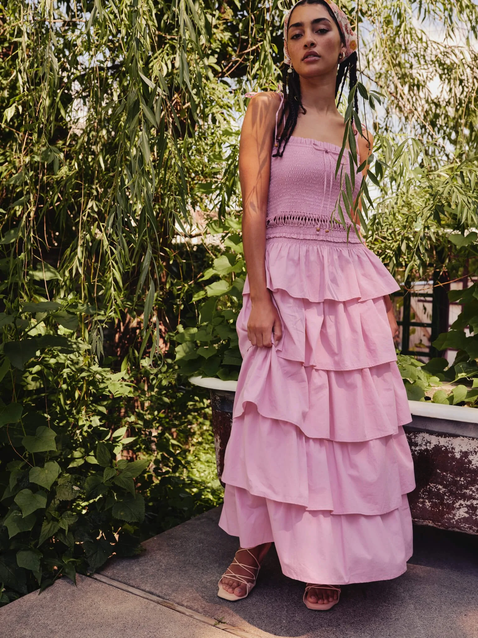 A woman in a pink layered dress standing outdoors among green foliage and trees.