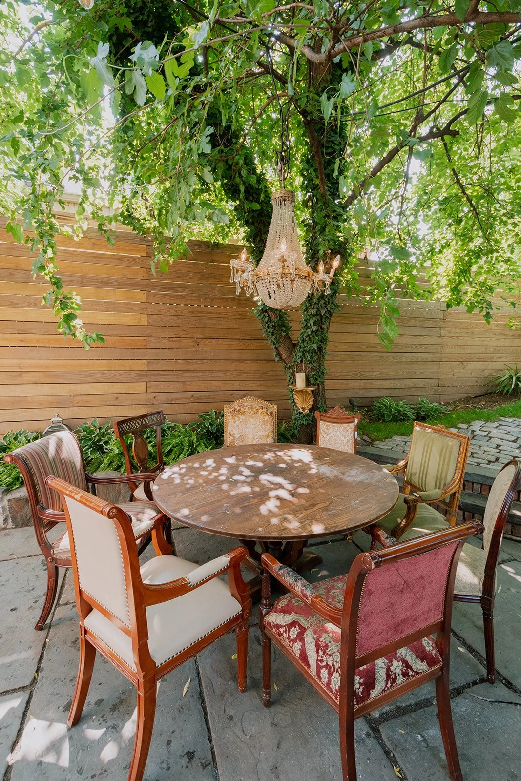An outdoor patio with a round wooden table surrounded by vintage upholstered chairs. A chandelier hangs from a leafy tree, casting dappled sunlight on the scene. A wooden fence and lush greenery are in the background.