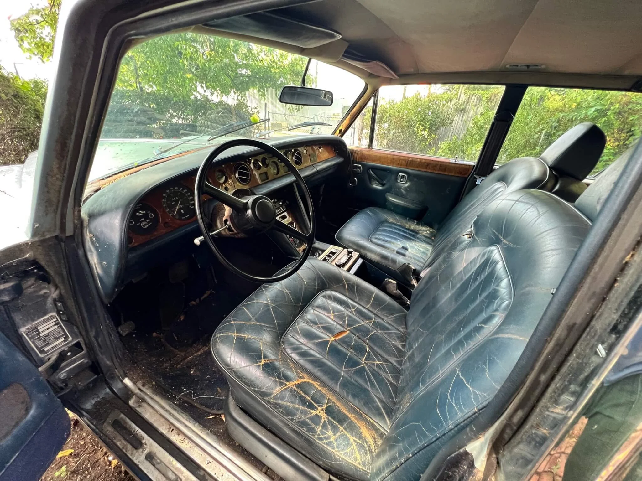 Interior of an old, worn-out car with black leather seats, a wooden dashboard, and a steering wheel. The driver's seat shows significant cracking and tears in the leather. The car is parked outdoors, with greenery visible through the windows.