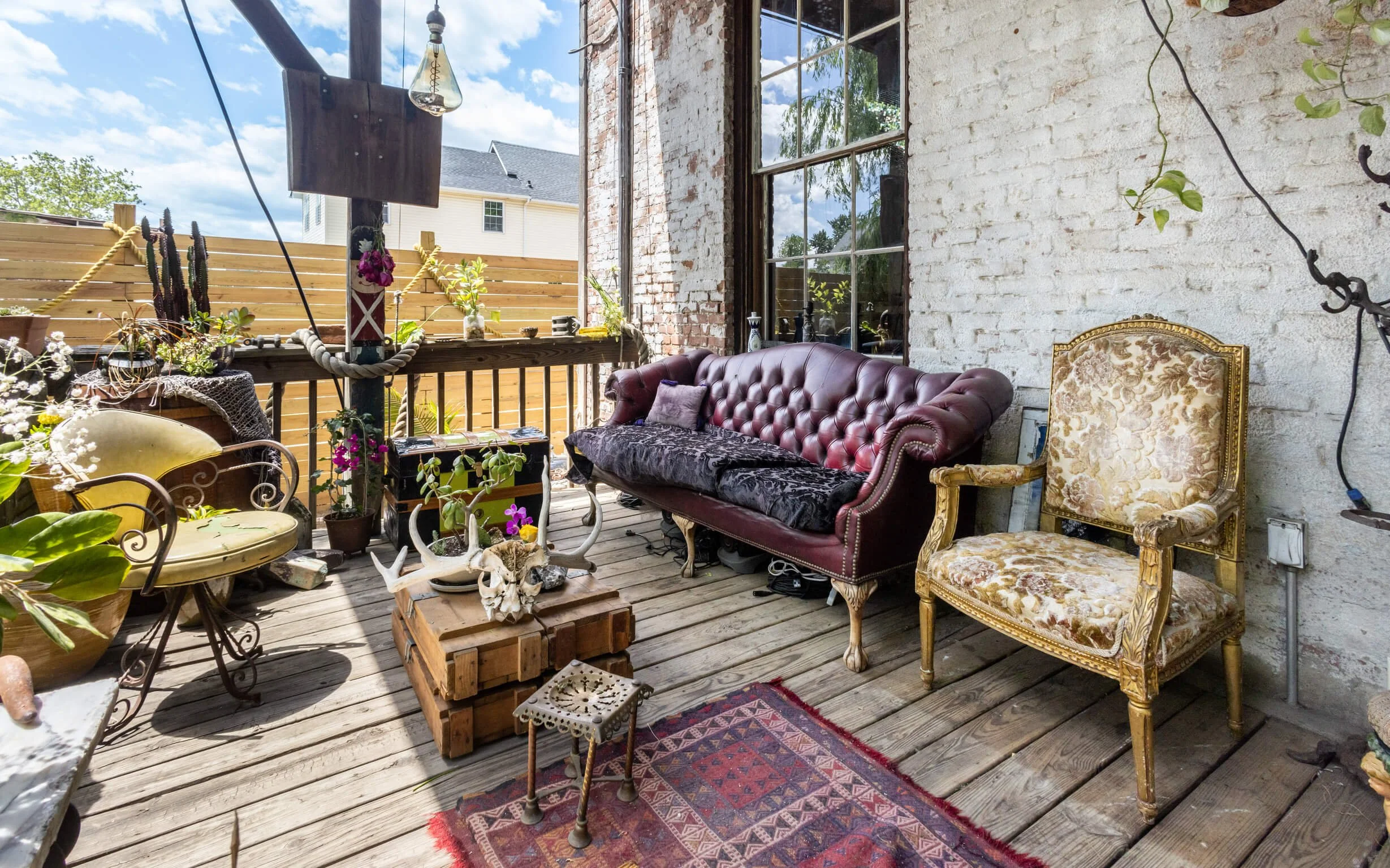 A rustic outdoor porch decorated with vintage furniture, potted plants, and eclectic ornaments. Features include a burgundy tufted leather sofa, a floral upholstered armchair, a small wooden table, a nested side table, and a colorful rug on weathered
