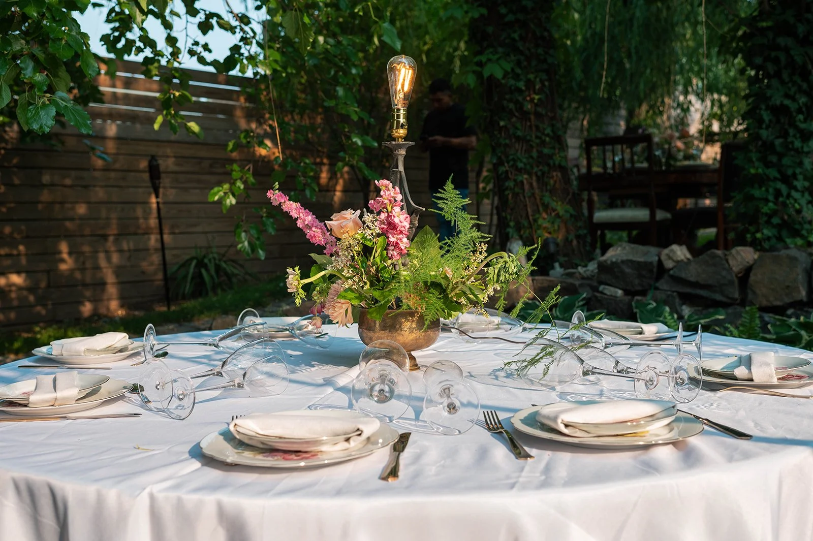 A round outdoor dining table with a white tablecloth, decorated with a central floral arrangement and a modern light bulb centerpiece. The table is set with white plates, folded napkins, and silverware, with wine glasses placed upside down. The backg