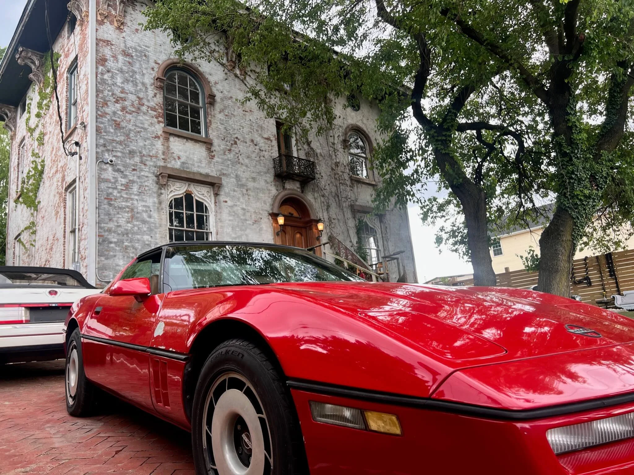 A red sports car parked in front of an old, weathered brick house with arched windows and a small balcony, surrounded by trees.
