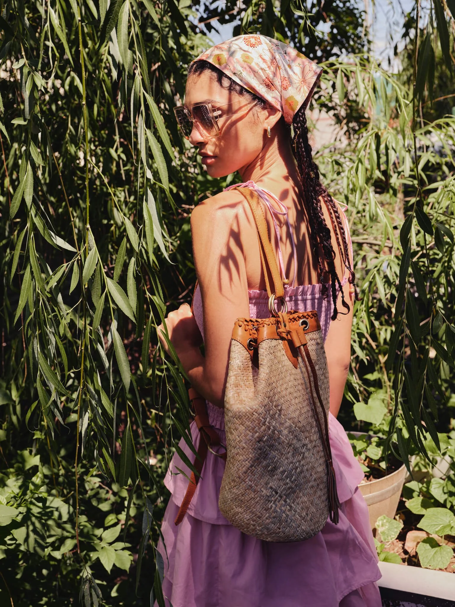 A woman with braided hair, wearing sunglasses, a floral headscarf, and a pink dress, standing among green foliage and trees.