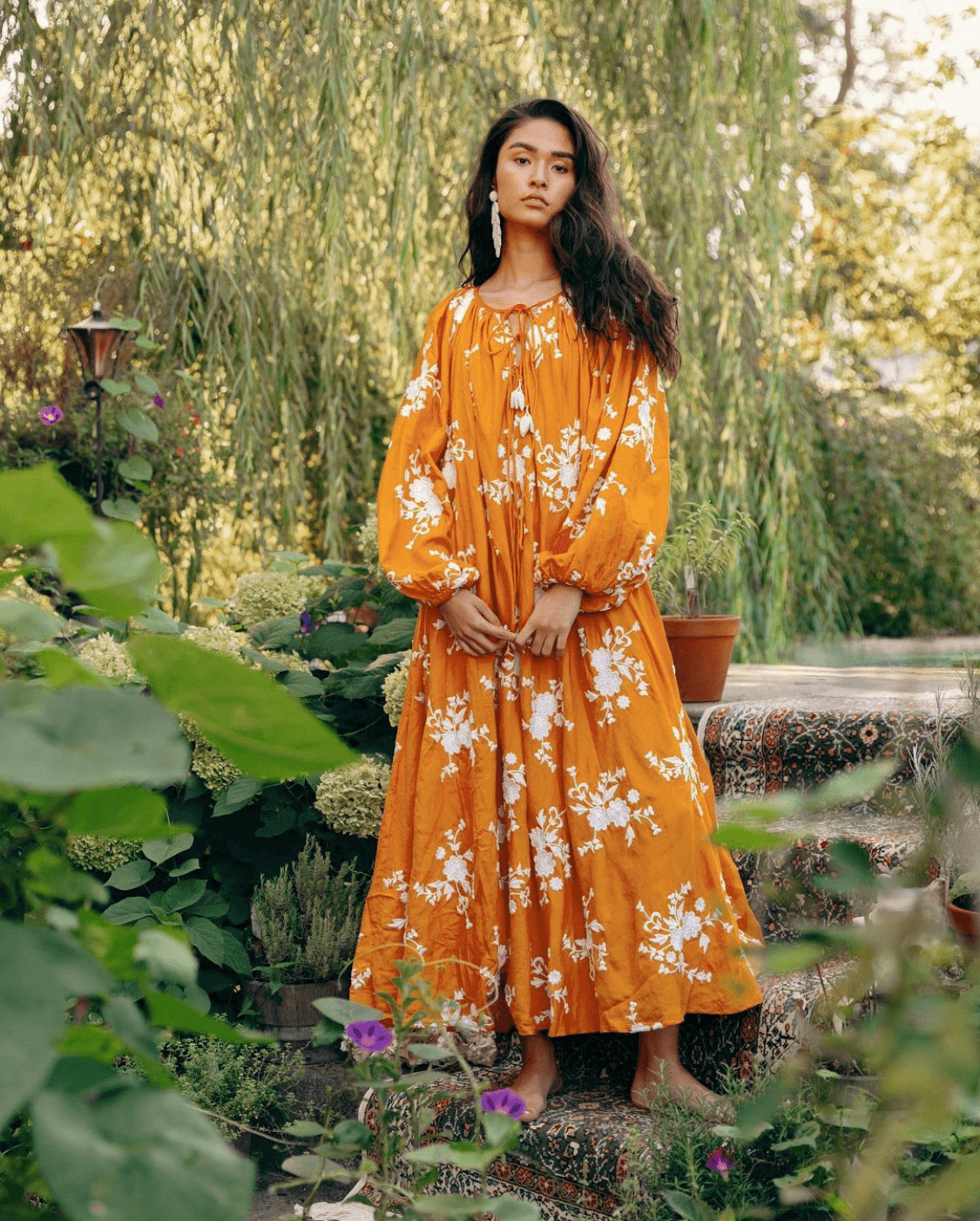 A woman with long dark hair wearing a flowing orange dress with white floral patterns, standing barefoot on a patterned rug outdoors surrounded by greenery and potted plants.
