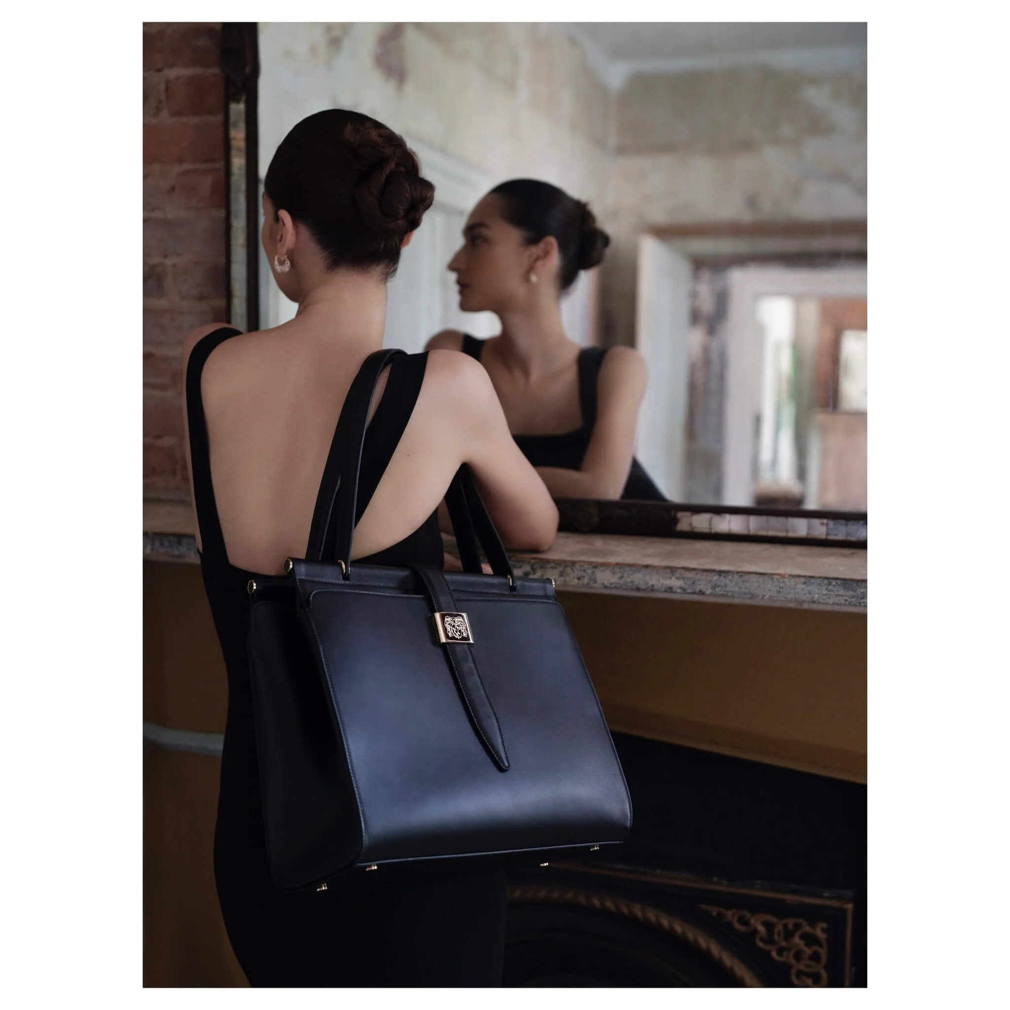 A woman with dark hair styled in an elegant updo looking at herself in a mirror. She is wearing a black sleeveless dress and has a large black leather handbag hanging from her shoulder. The setting appears to be a vintage-style room with exposed bric