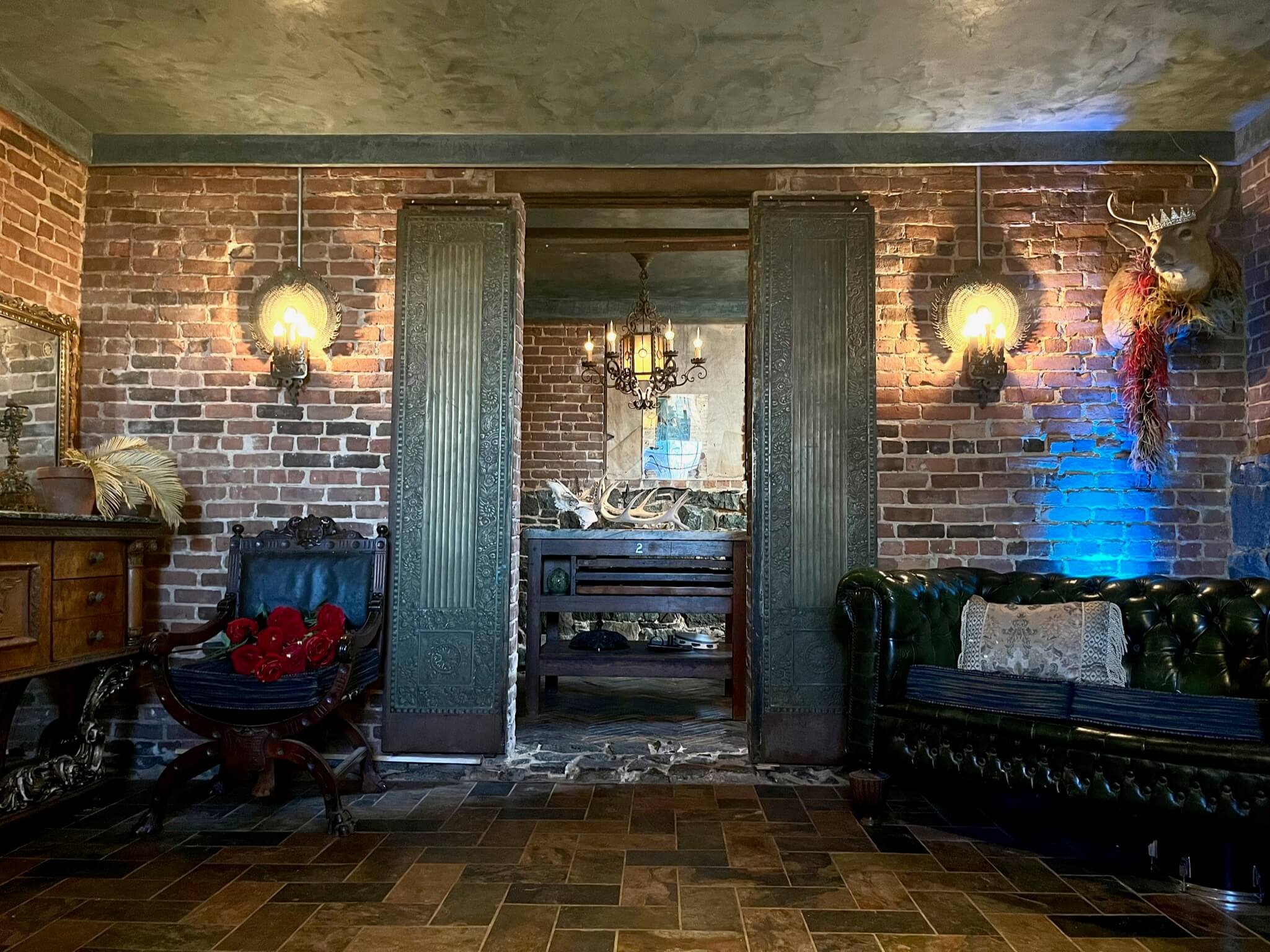 A rustic interior living space with exposed brick walls, antique furniture, and decorative elements such as a mounted deer head, feather arrangements, and warm lighting fixtures.