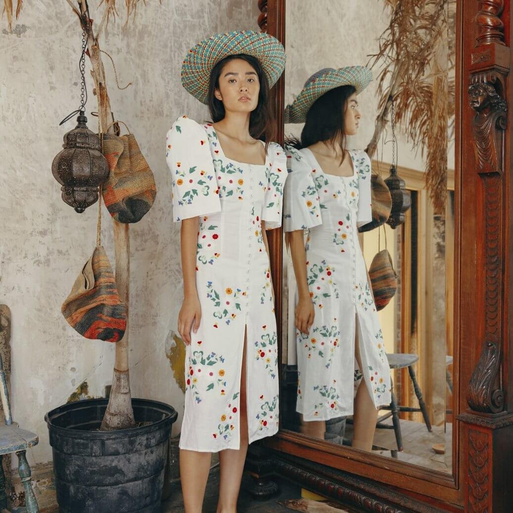 A woman in a white dress with colorful floral embroidery stands next to a mirror, wearing a large woven straw hat. The scene is set in a rustic room with vintage decor, including hanging woven bags and an antique chair. Her reflection is visible in t