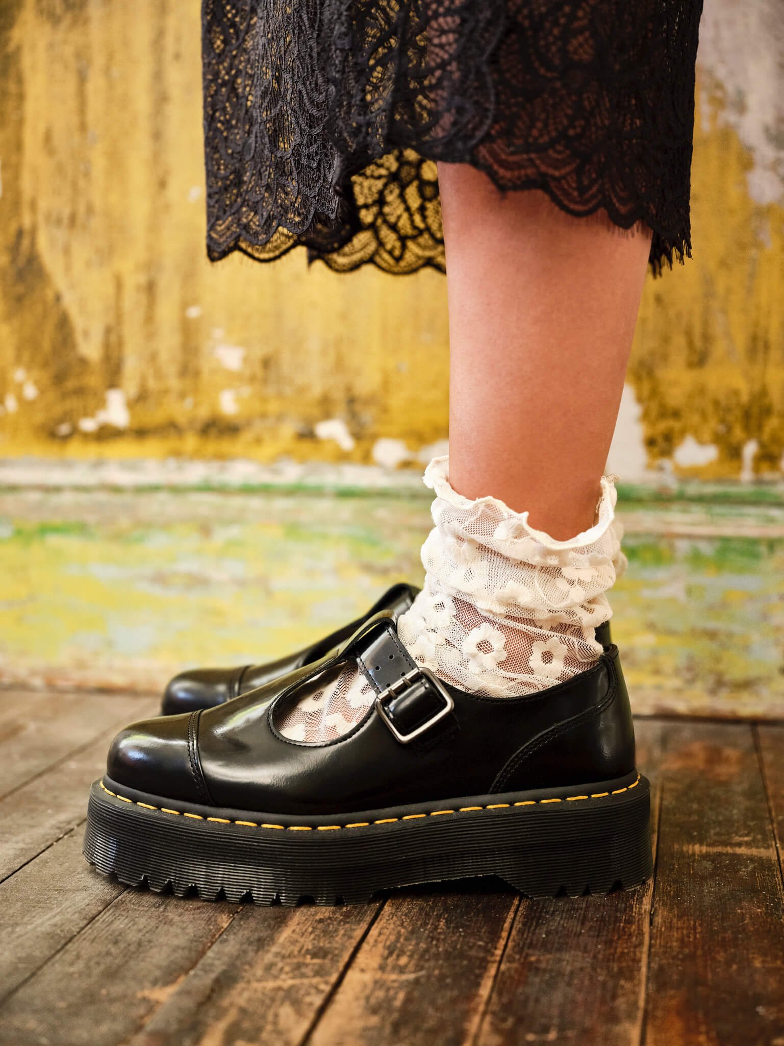 A close-up of a person’s lower leg wearing lace socks with a floral pattern, black platform Mary Jane shoes with buckle straps, and a black lace skirt, standing on a wooden floor with a yellow and green weathered wall in the background.