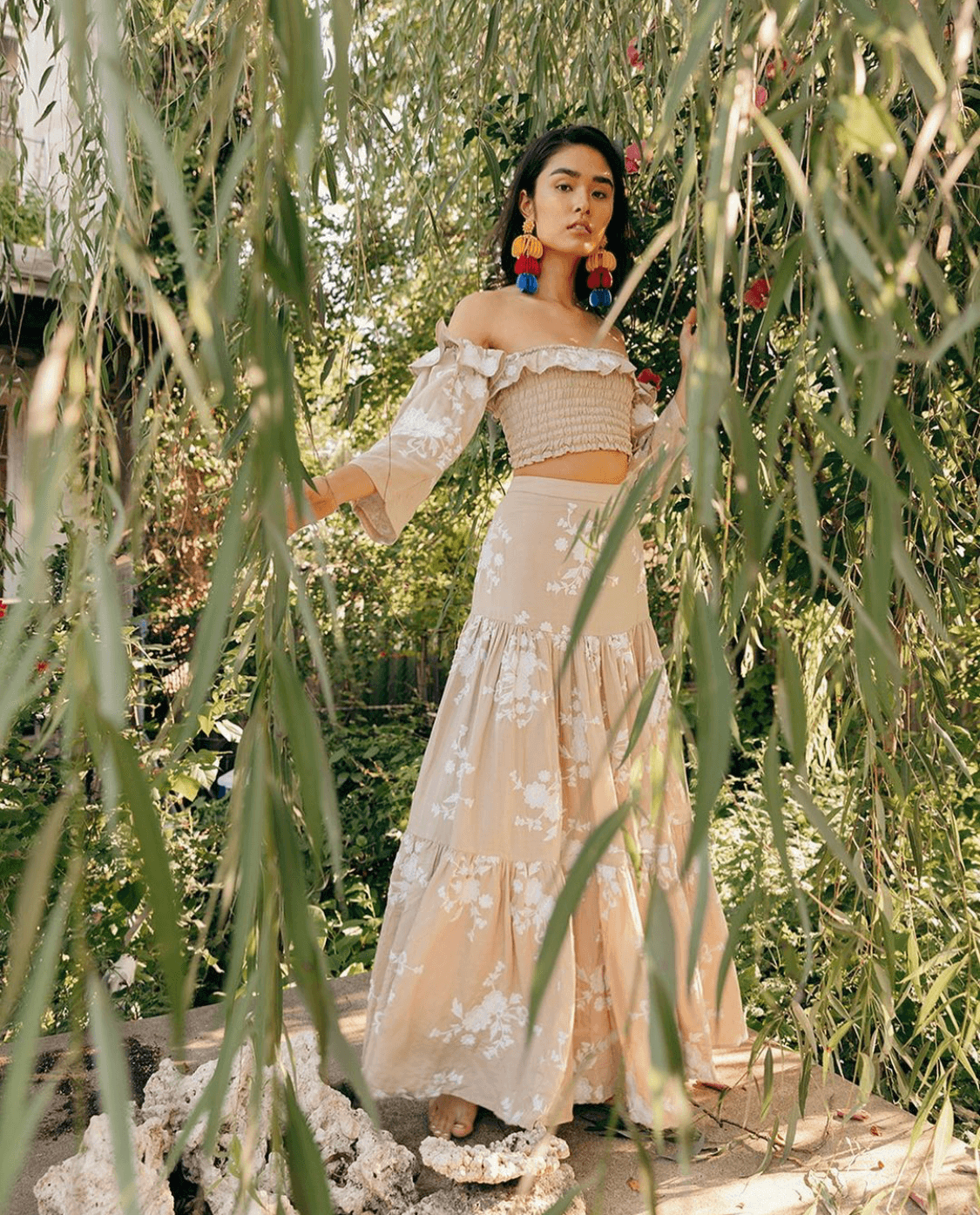 A woman standing among green foliage, wearing a beige off-shoulder ruffled crop top and a long beige skirt with white floral patterns, accessorized with colorful pom-pom earrings.