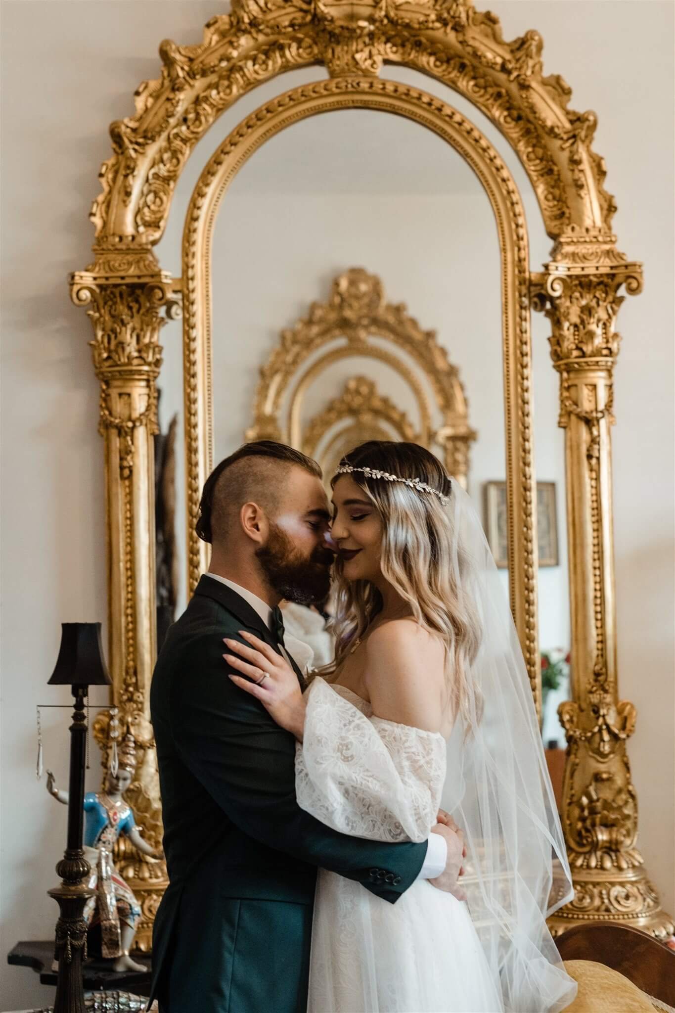 A bride and groom in wedding attire sharing a romantic moment indoors, standing in front of an ornate, gold-trimmed mirror, with wedding details visible in the background.