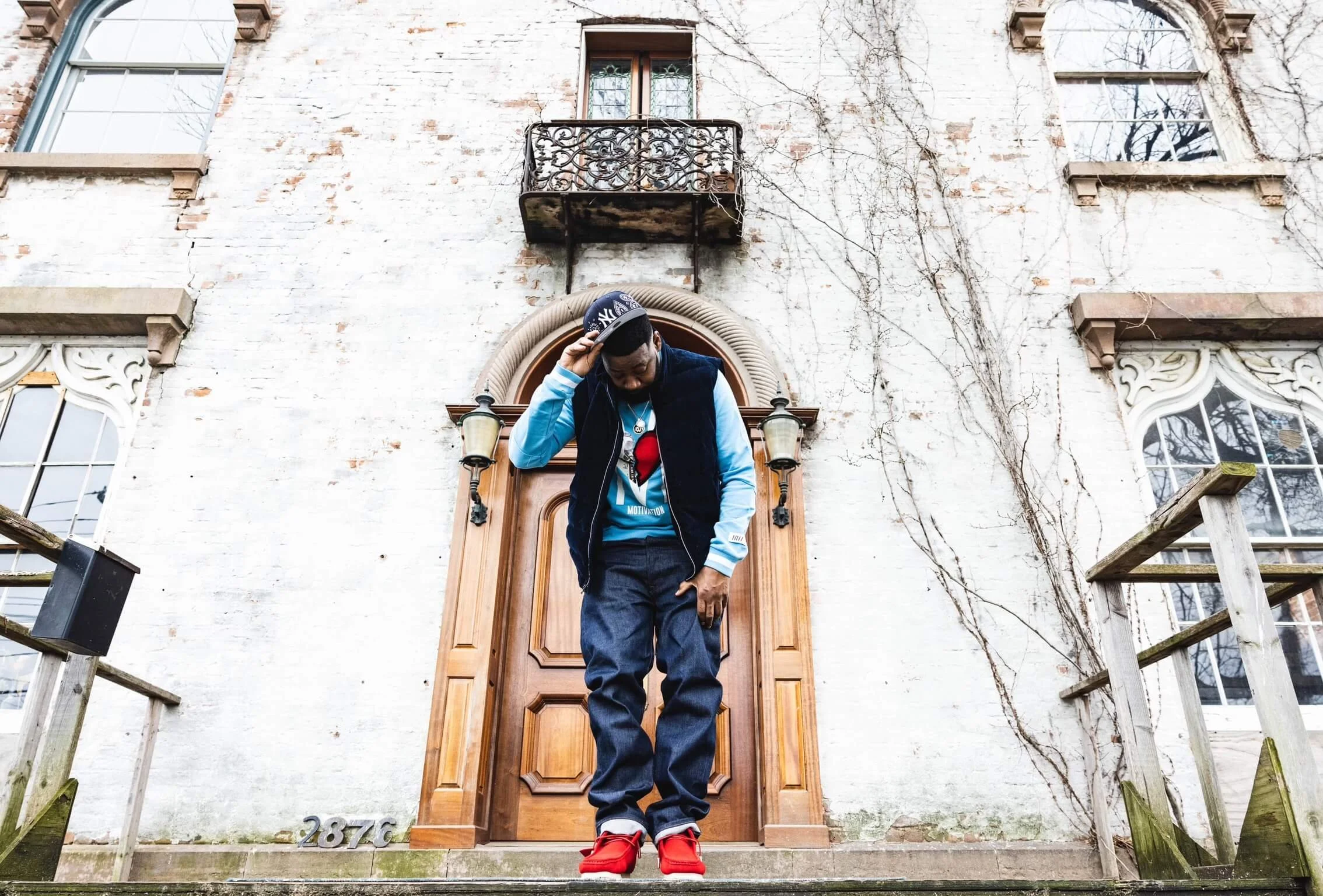 Young man standing on front steps of a house, dressed in casual streetwear, adjusting his cap, with a brick building exterior and vintage-style balcony above.