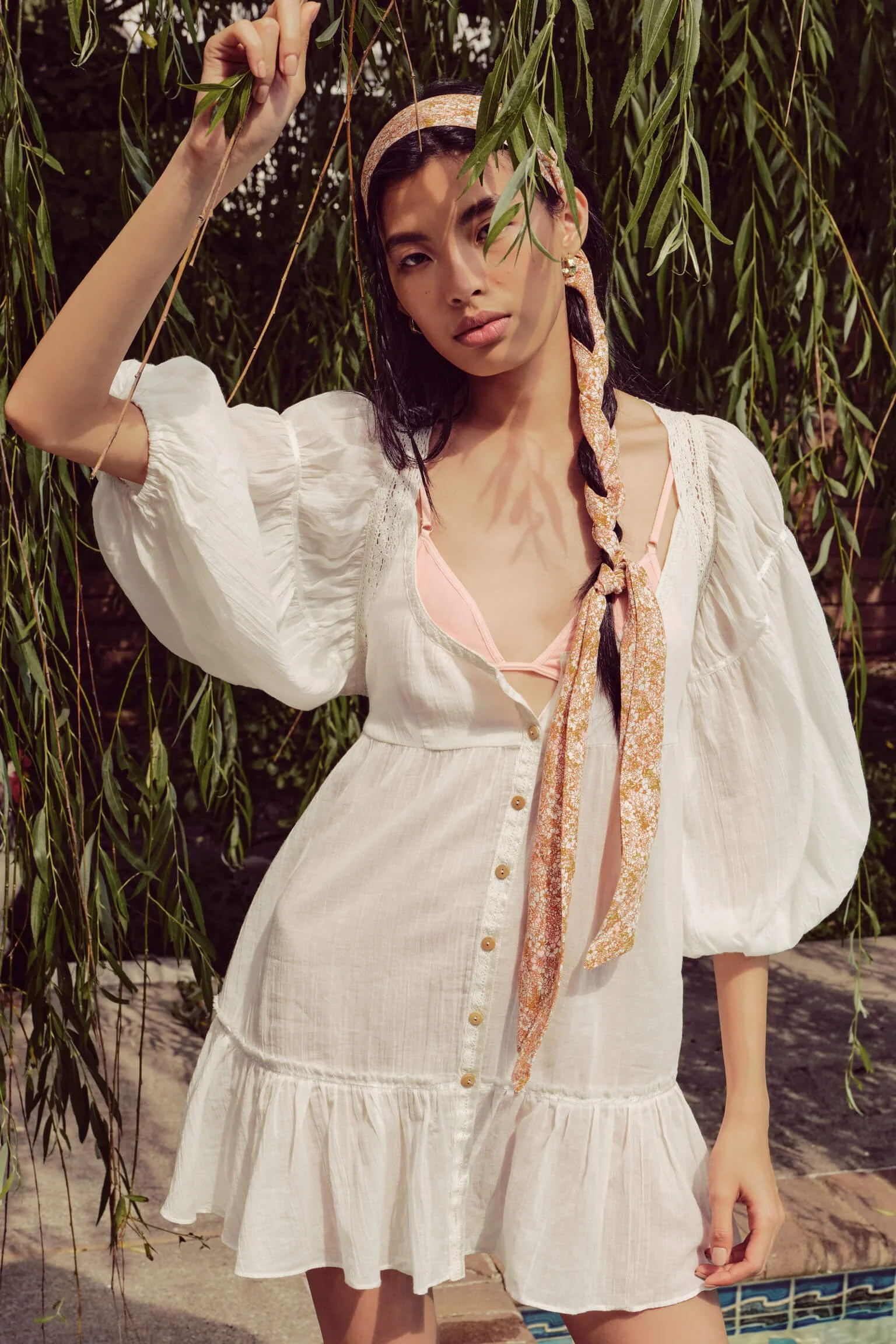 A woman standing outdoors among hanging green foliage, wearing a white dress with puffy sleeves, buttons down the front, and a ruffled hem. She has a patterned scarf tied around her neck, a headband, and is holding a branch with leaves.