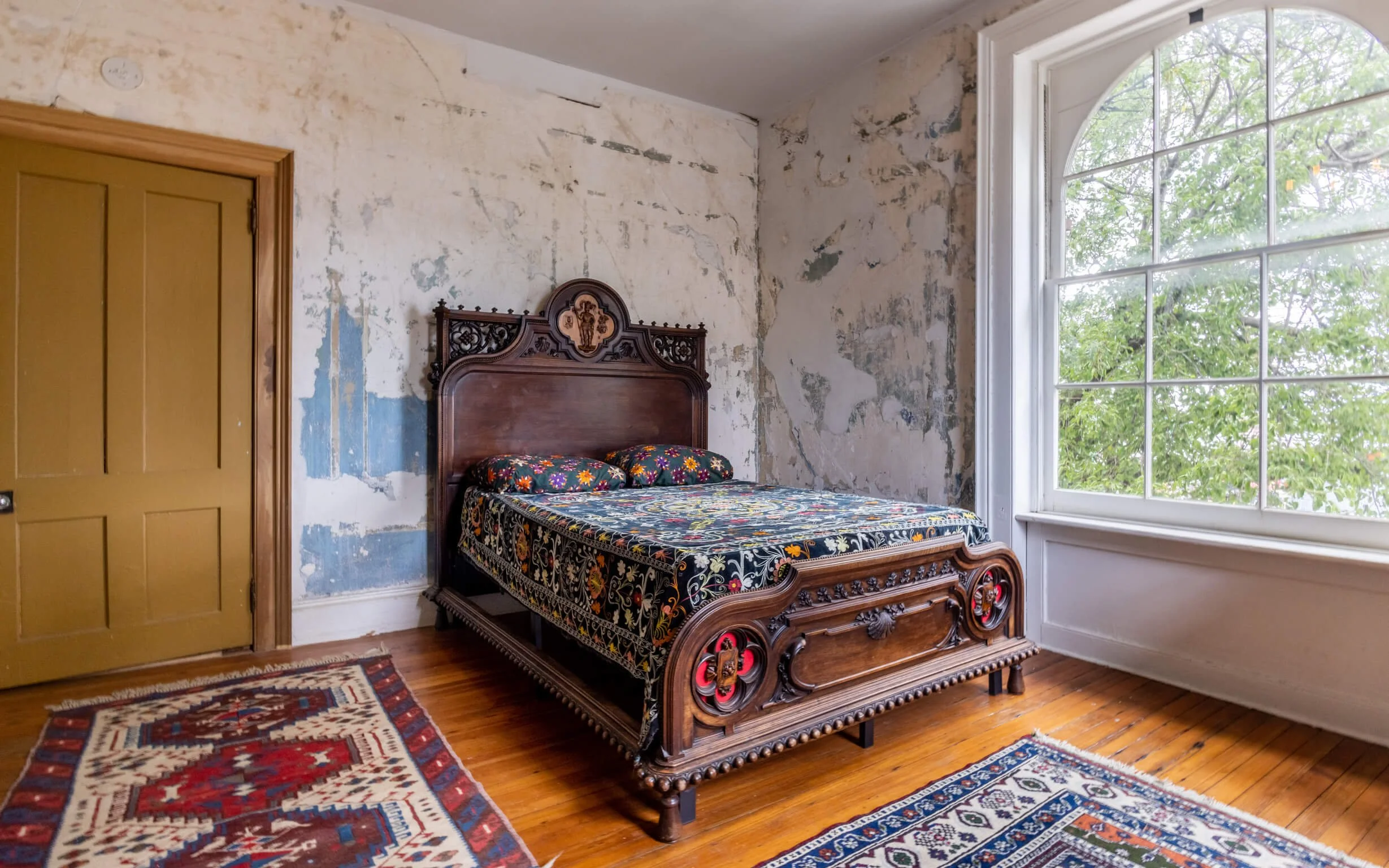 Bedroom with wooden bed, floral quilt, worn wall with peeling paint, large window, and oriental rugs on hardwood floor.