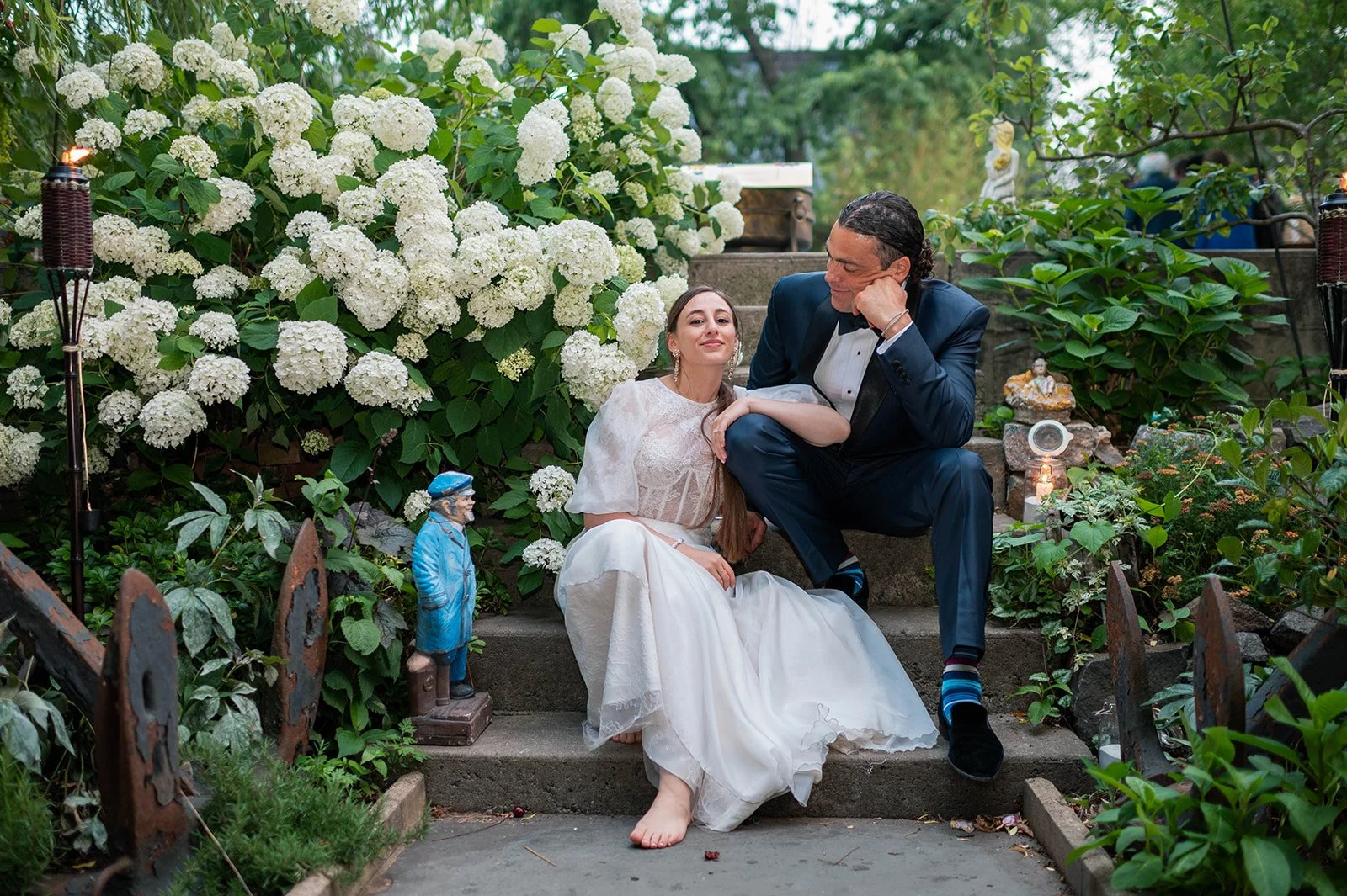A bride and groom sitting on garden stairs surrounded by white hydrangea flowers and greenery, with decorative garden statues and candles nearby.
