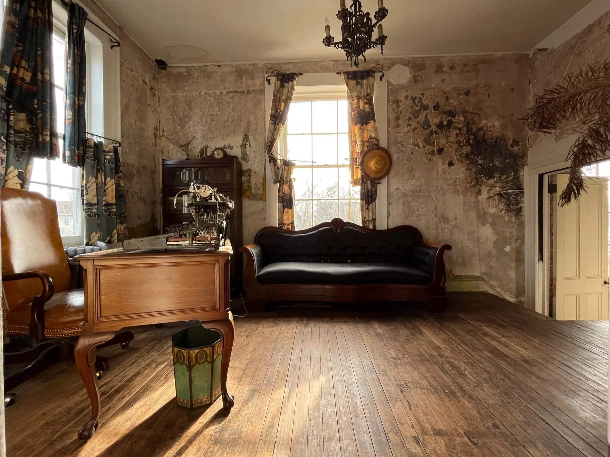 Vintage room with worn, peeling walls, large windows with patterned curtains, wooden flooring, and antique furniture including a leather armchair, wooden desk, black sofa, and a cabinet. There's a chandelier hanging from the ceiling and a hat on the 