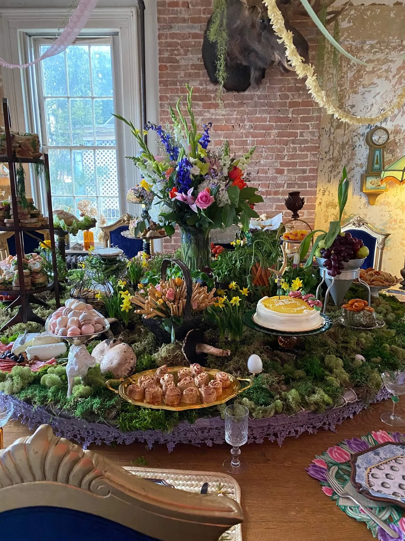 A decorated dining table with an outdoor-themed centerpiece of flowers, moss, and moss-like decorations, with various desserts and foods displayed, including a cake, cookies, and grapes, in a room with brick walls and a large window.