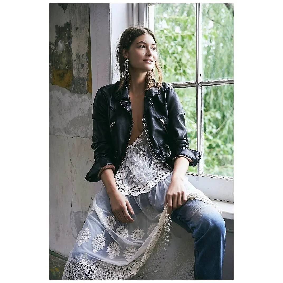 A woman sitting by a window, wearing a black leather jacket over a lace dress, with greenery outside.
