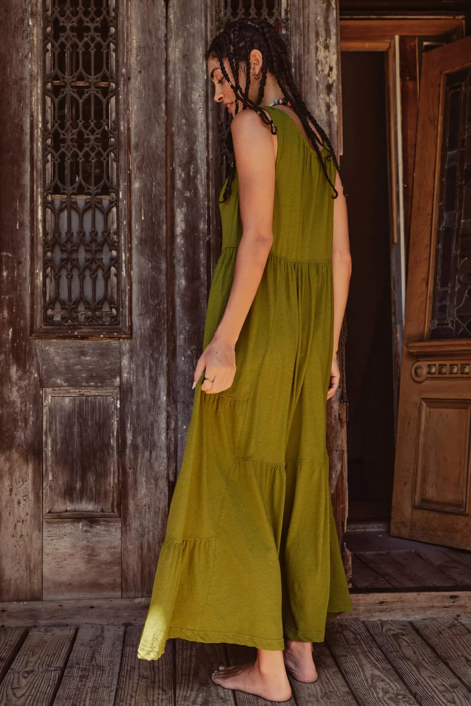 A woman with long braided hair wearing a loose, green maxi dress, standing barefoot on a wooden floor in front of a rustic wooden door and wall.