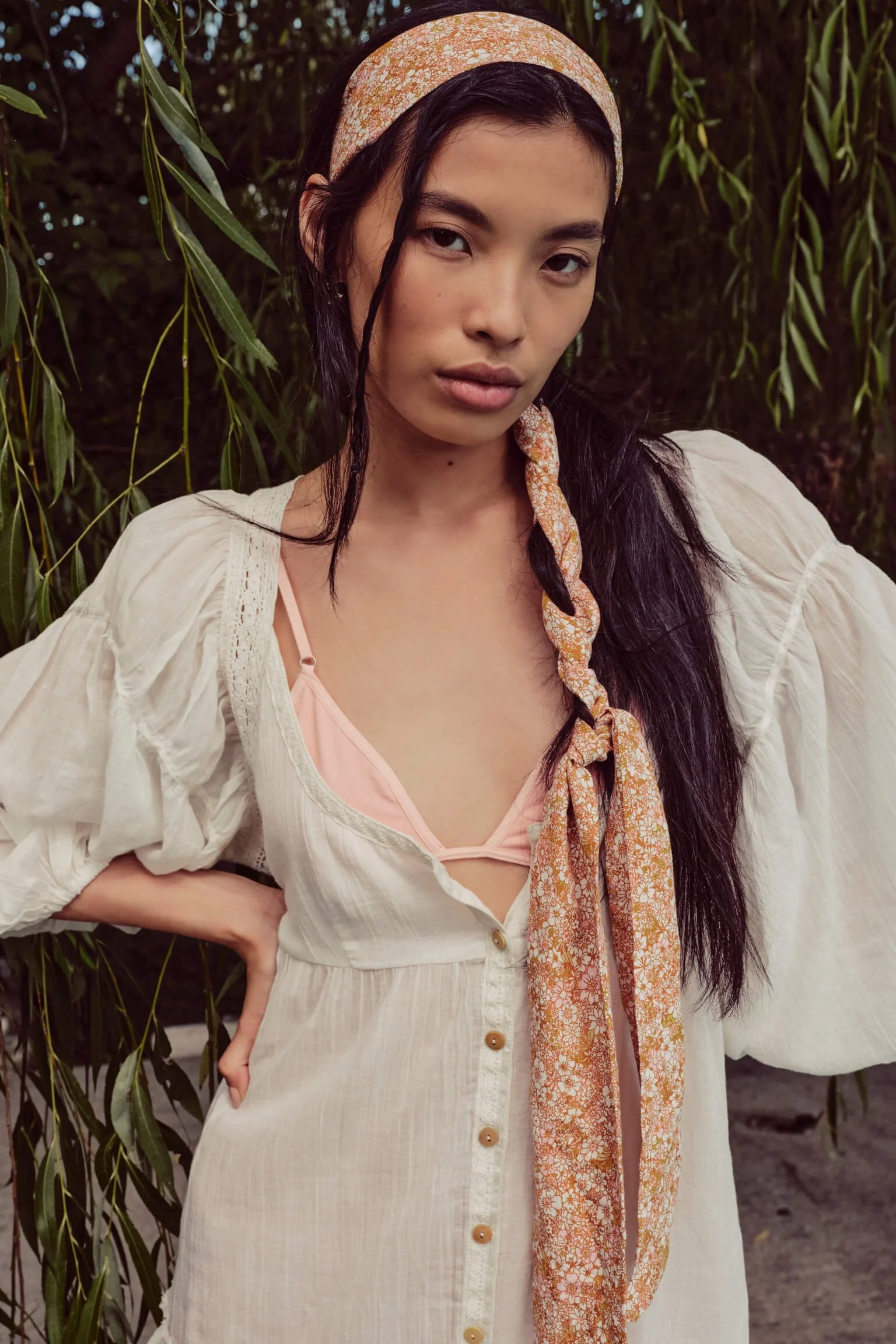 A young woman with long dark hair, wearing a peach headscarf and a white dress with puffy sleeves, standing outdoors among green foliage, looking at the camera.