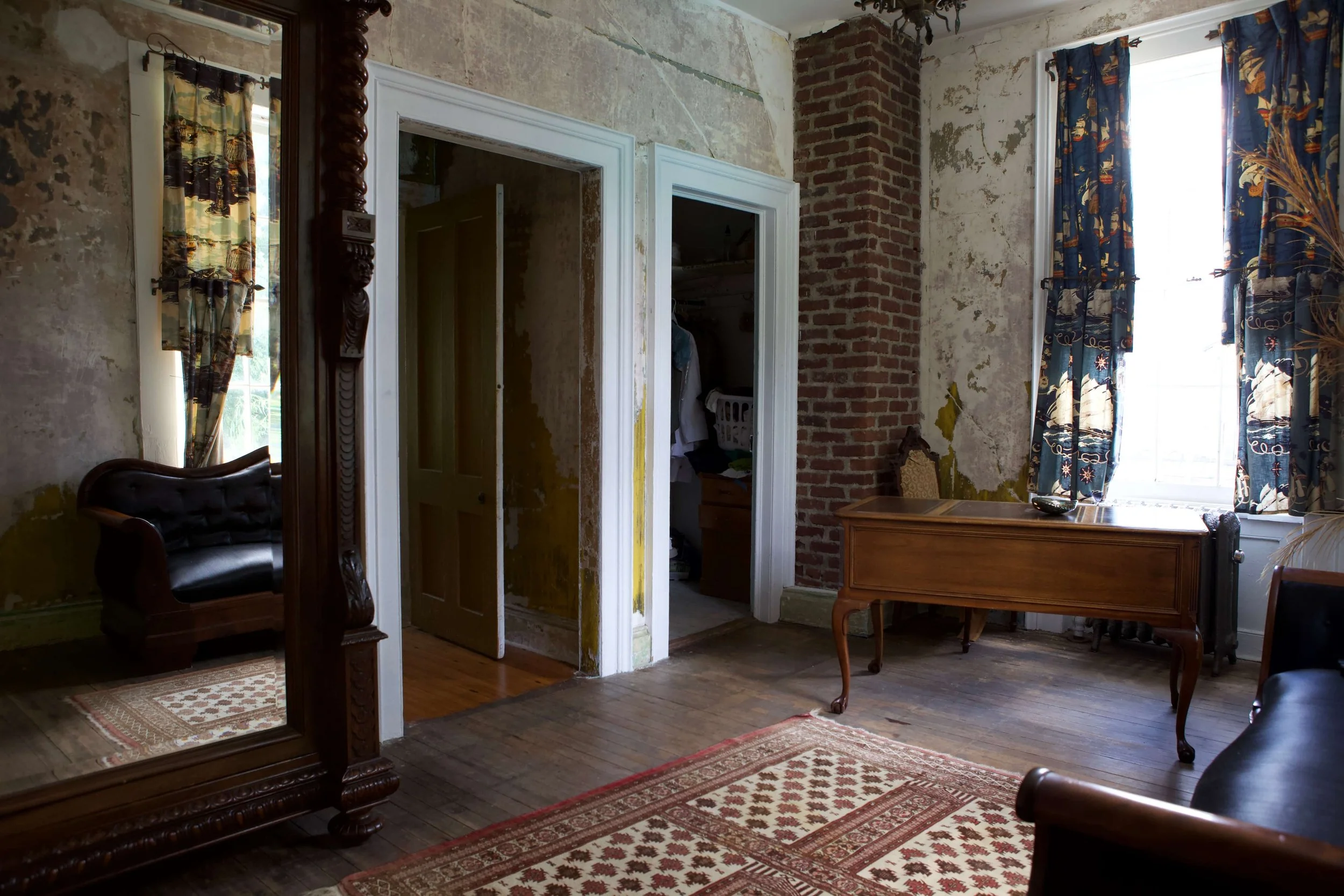 Interior of a vintage-style room with worn walls, a rug, a wooden table, and a large mirror reflecting part of the room. Open doorway revealing a closet and another doorway leading to a space with clothes.