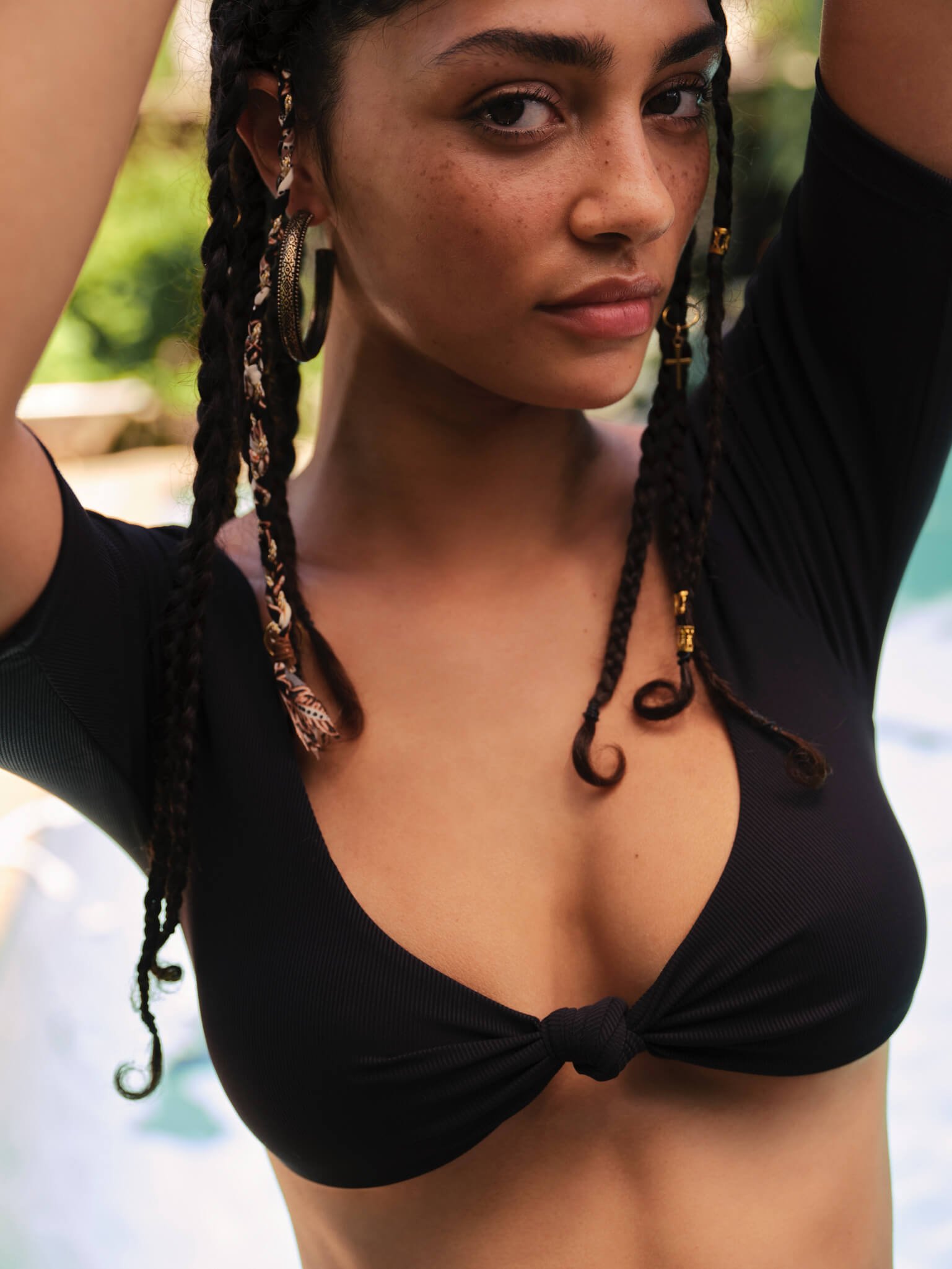 Close-up of a young woman with braided hair, wearing large hoop earrings and a black top with a knot at the center, outdoors with a blurred background.