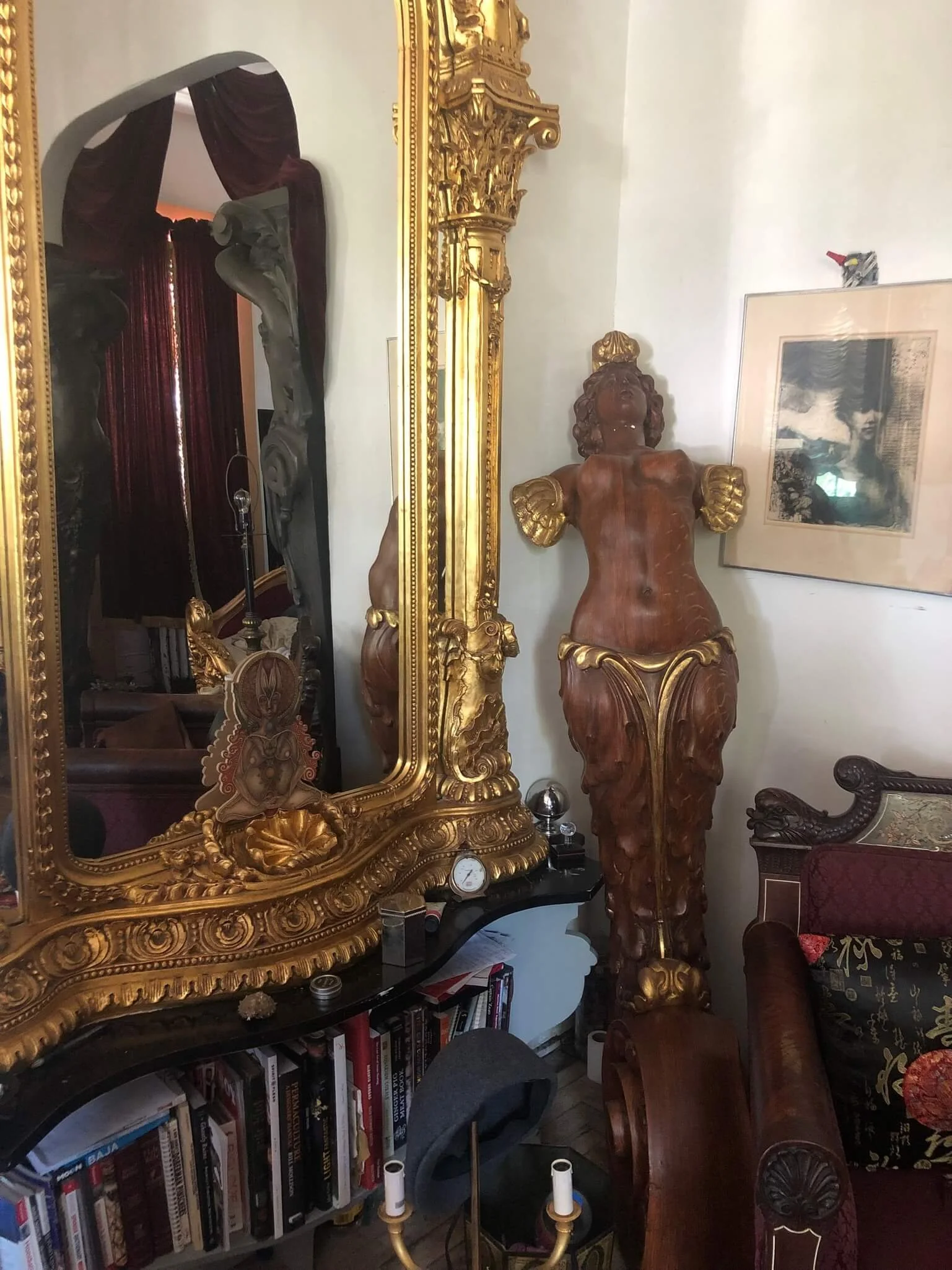 An ornately decorated room with a large gilded mirror, a wooden sculpture of a semi-nude woman with hair and gold details, a framed artwork on the wall, books on a shelf, and a vintage armchair with a cushion.