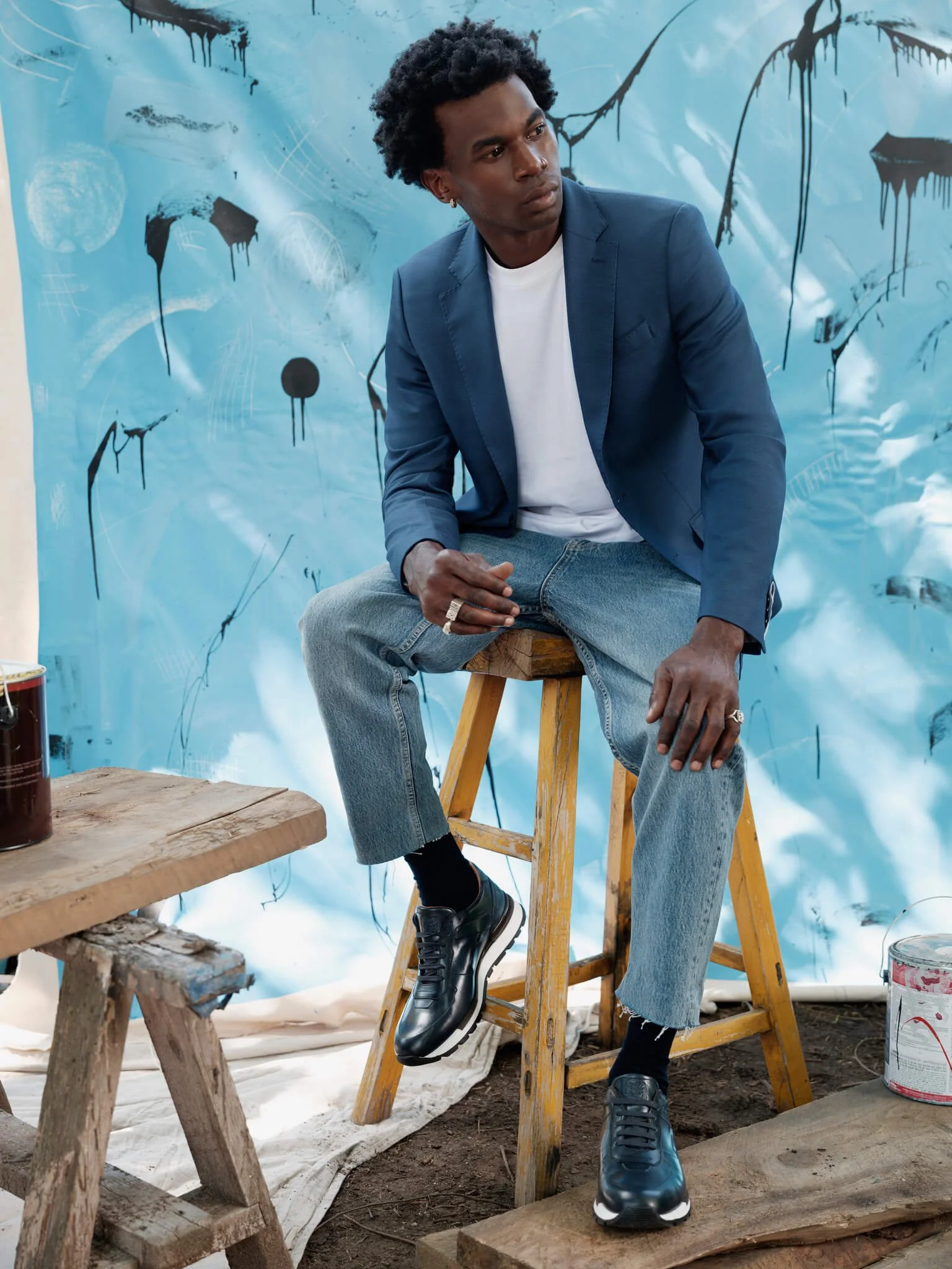 A young man with curly hair wearing a blue blazer, white t-shirt, and jeans sitting on a wooden stool in front of a blue abstract painted backdrop. There are paint cans and a wooden table nearby.