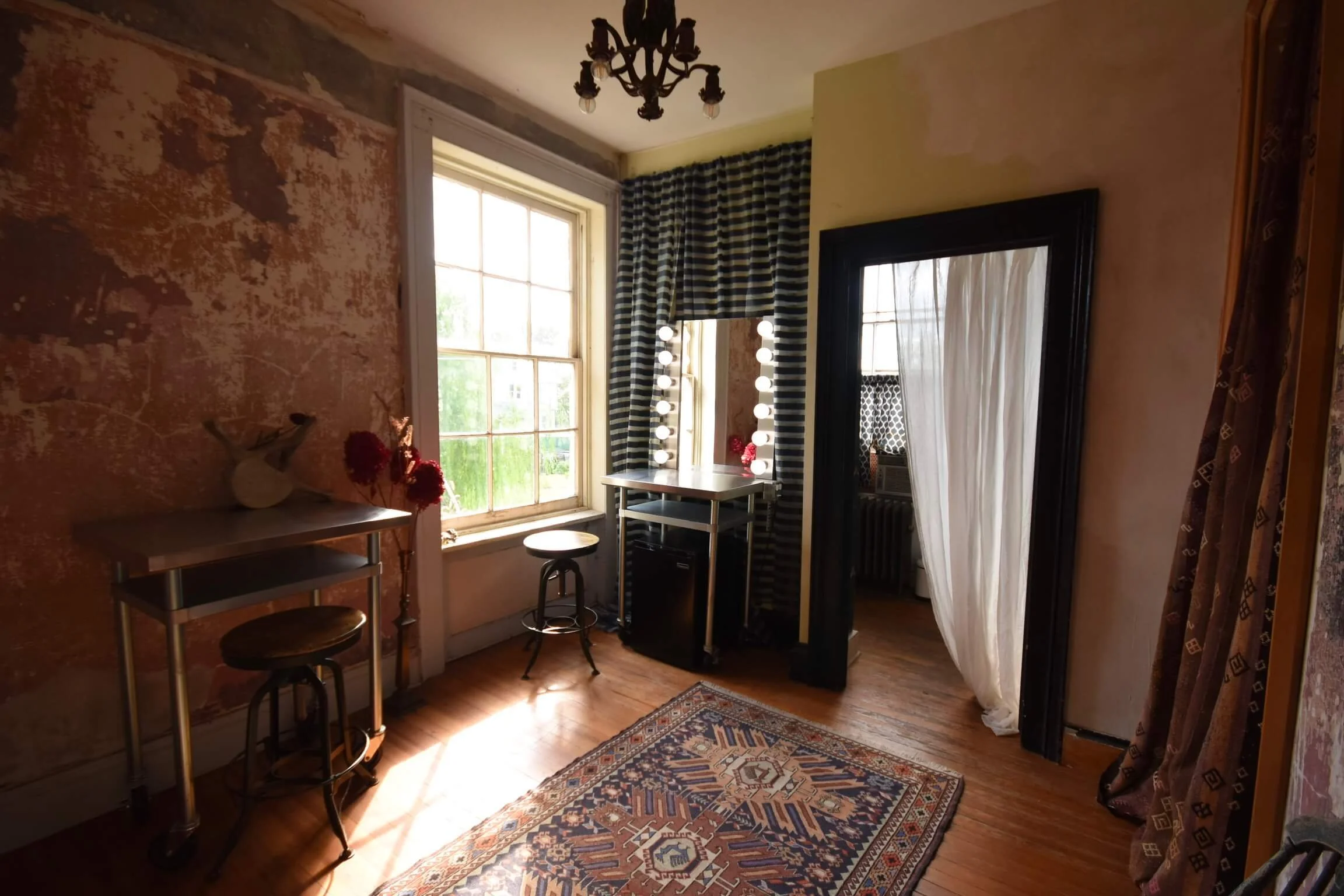 A vintage-style room with a floral rug, a window with black and white striped curtains, and a vanity mirror with lights. There is a table with a deer head sculpture and a vase of red flowers, and another small table with two stools.