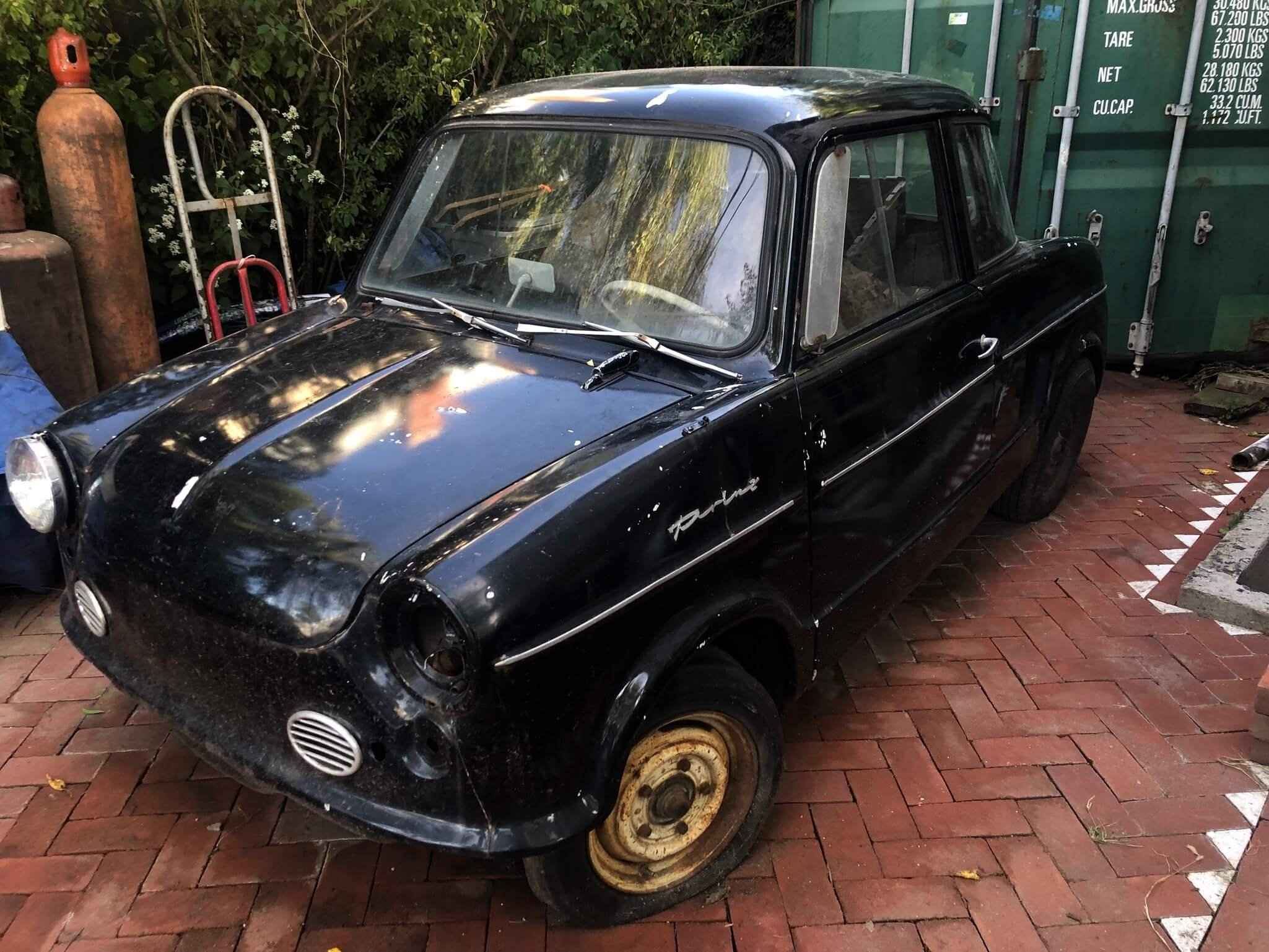 Old black compact car with a rusted wheel, parked on a brick driveway next to gas cylinders and greenery.