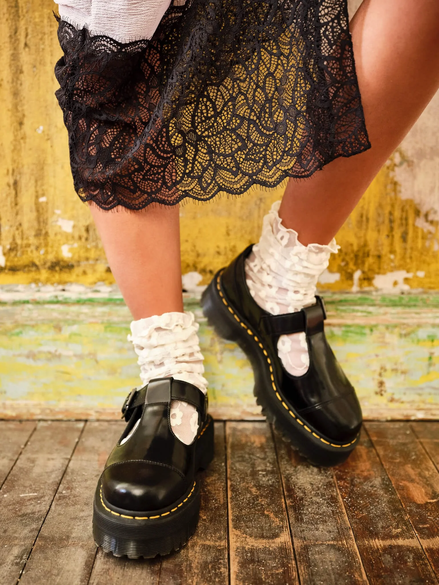 Close-up of a person's outfit, showing black lace skirt, white floral socks, and black platform Dr. Martens shoes, standing on a wooden floor with a peeling yellow and white wall in the background.