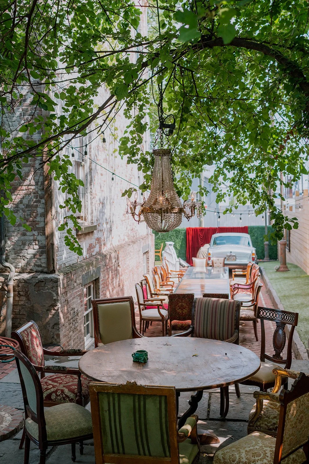 An outdoor patio with vintage chairs, a large wooden table, and a decorative chandelier hanging from tree branches.