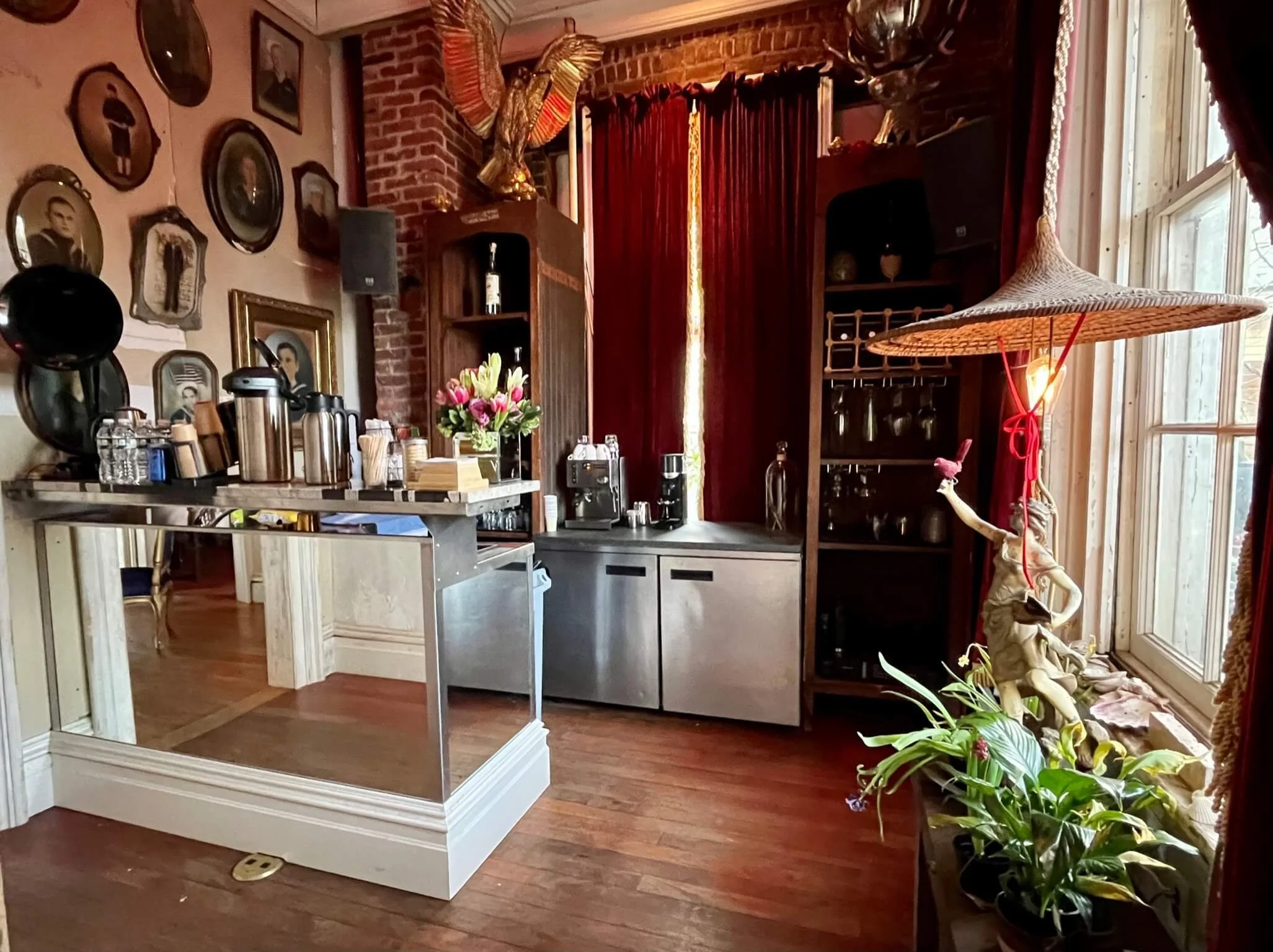 A cozy interior with a bar counter, framed vintage photos on the wall, a bouquet of flowers, and a bamboo lamp near a window with red curtains.