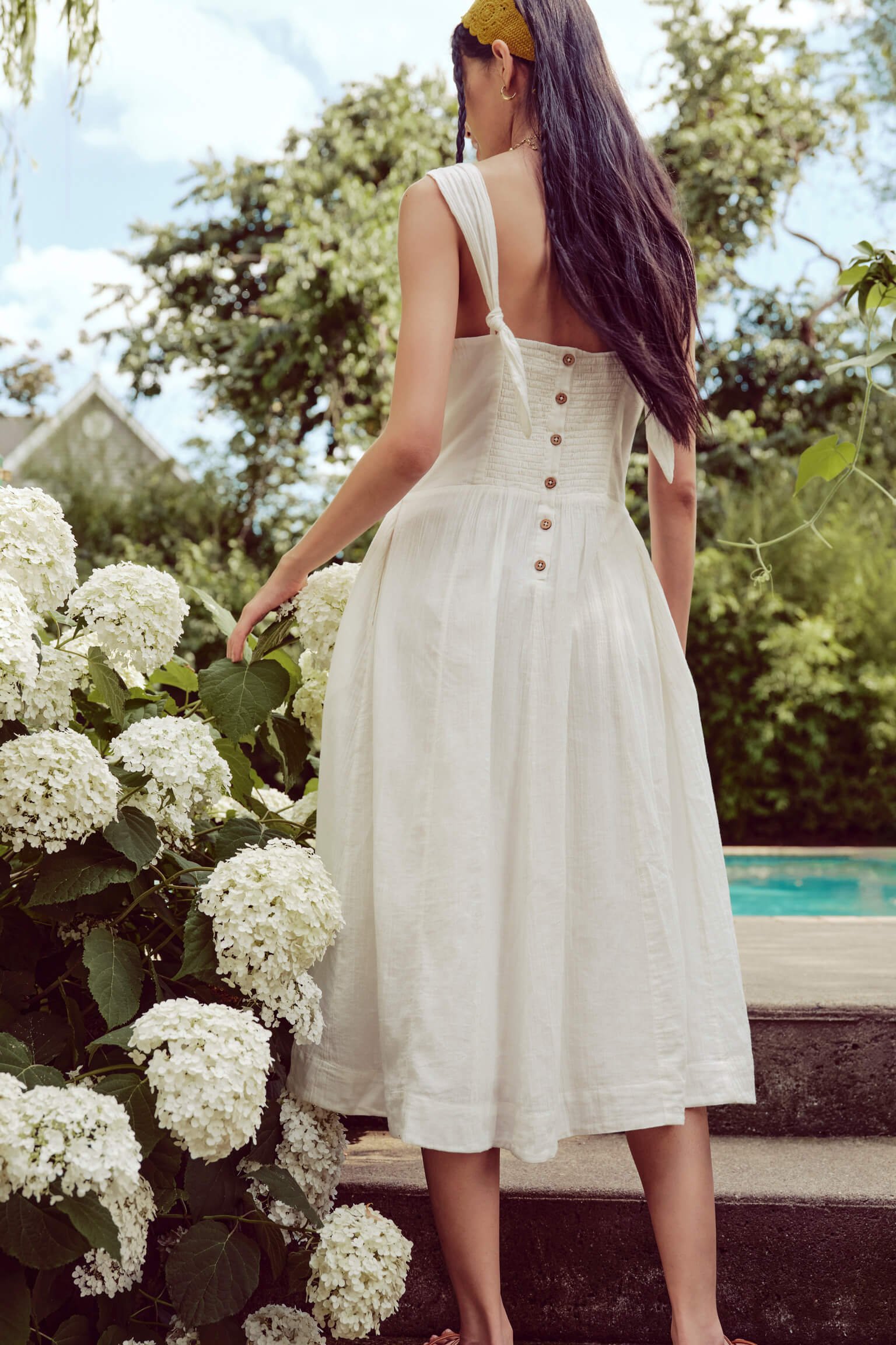 Woman in a white sleeveless dress with straps and wooden buttons, standing outdoors near white hydrangea flowers, with greenery and a swimming pool in the background.