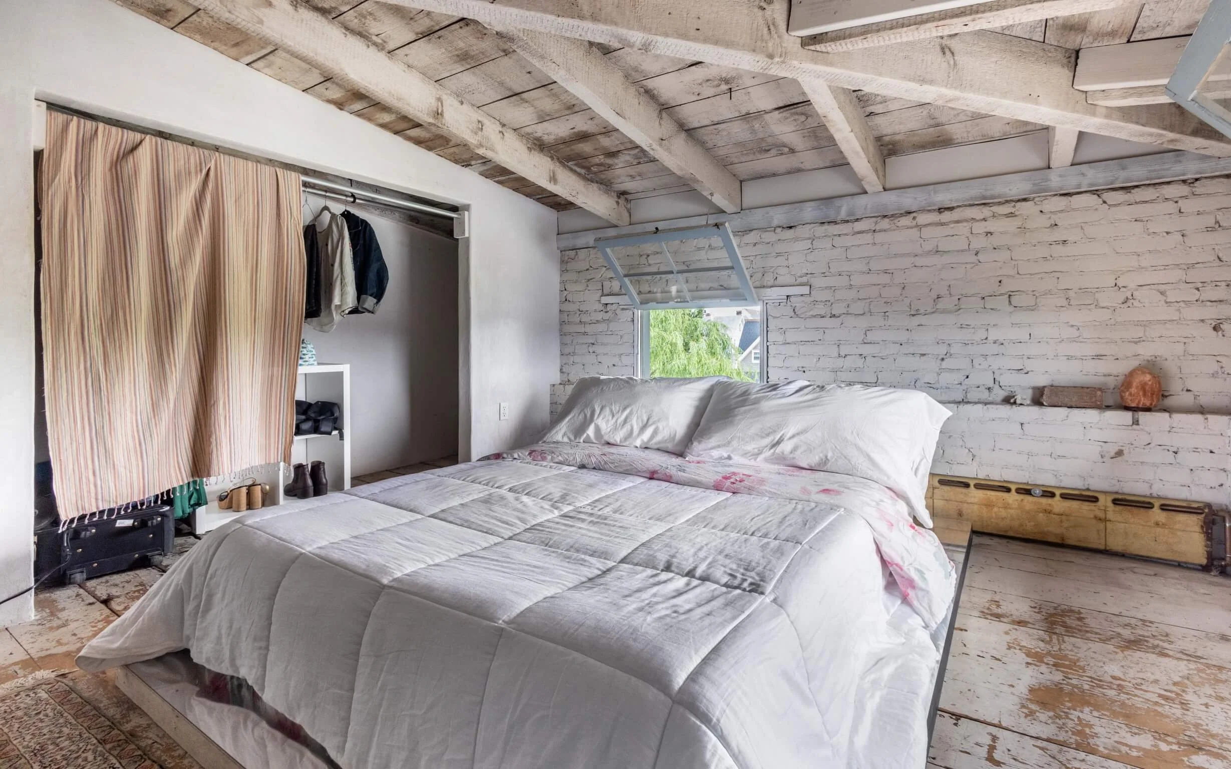 A rustic bedroom with white brick walls, exposed wooden beams, and a boarded-up window. There is a bed with white bedding and an open window above it. To the left, a small closet area with a clothes rod, a pink curtain, and shoes on the floor. The fl