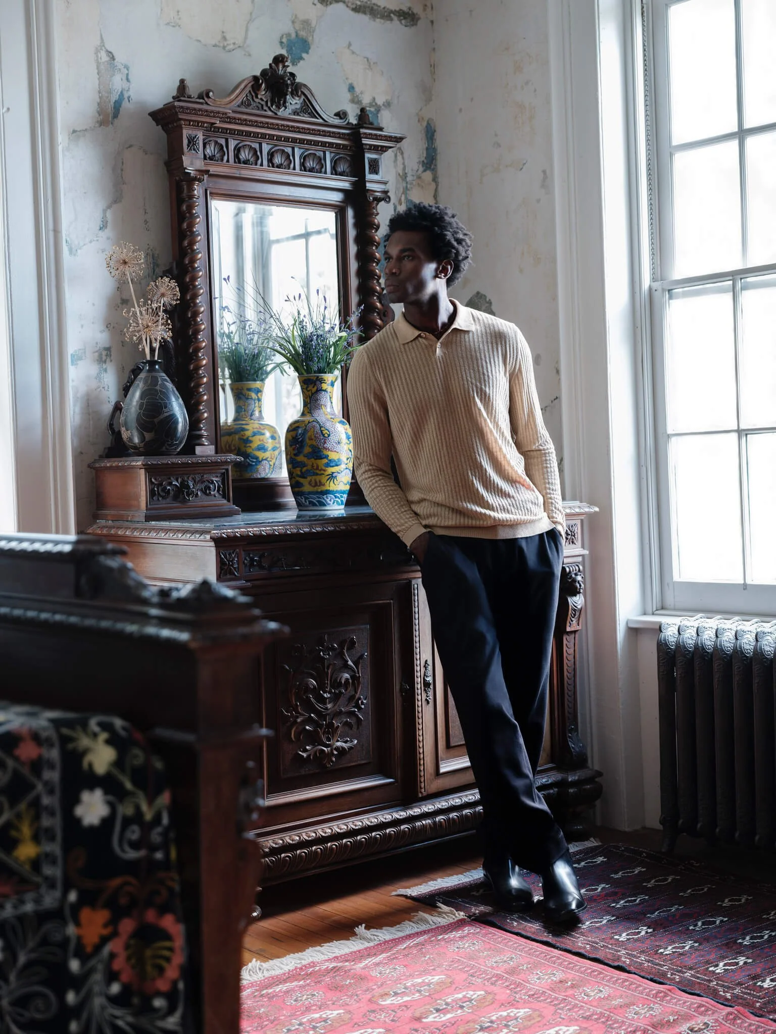 A man with dark skin and curly hair leaning against a vintage wooden dresser with a mirror in a room with peeling paint walls and a large window. He is wearing a cream-colored ribbed sweater, black pants, and black shoes. There are decorative vases w