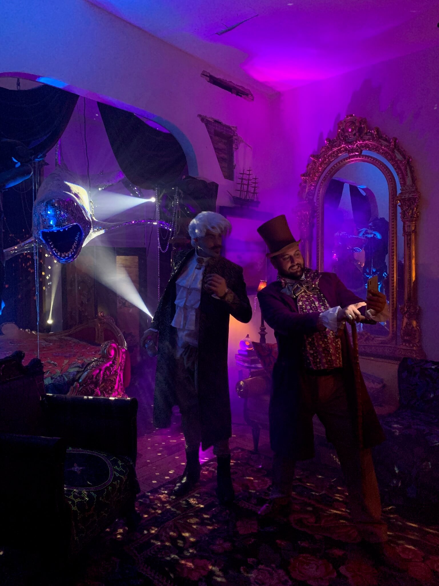 Two men dressed in elaborate costumes stand in a dimly lit, colorful room with purple and pink lighting. One is wearing a tall top hat and ornate clothing, taking a selfie, while the other, dressed in period attire, is looking at him. The room has a 