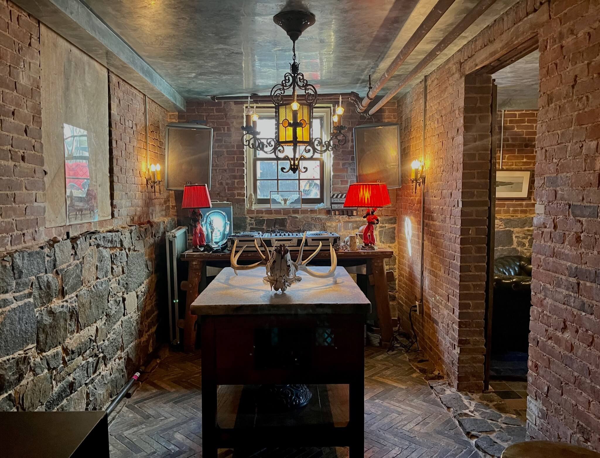 A rustic room with exposed brick walls, a stone wall, and wooden floors. There is a wooden table in the center with antlers and a skull on top. Two red table lamps with red shades flank the back wall, which has a window with a grid pattern. On the ba